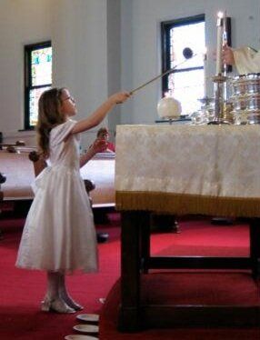 Girl in white dress lighting a candle at a church altar; red carpet, sunlight through windows.