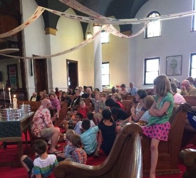 Children and adults in a church, gathered for an event. People are sitting on the pews, talking and watching.