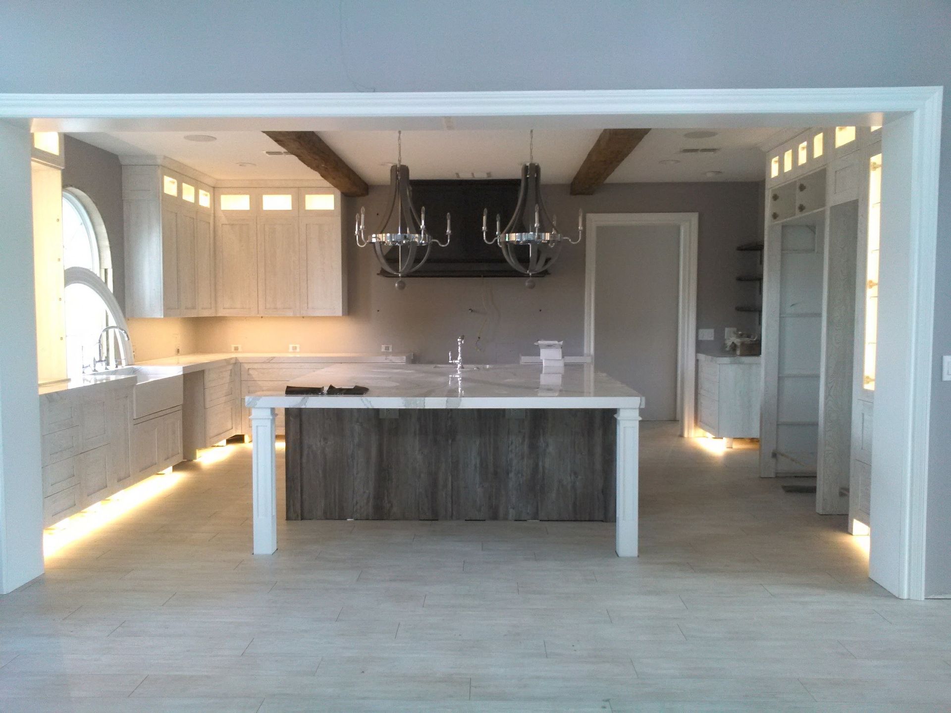 Kitchen with light wood cabinets, white island, two chandeliers, and wooden beams.