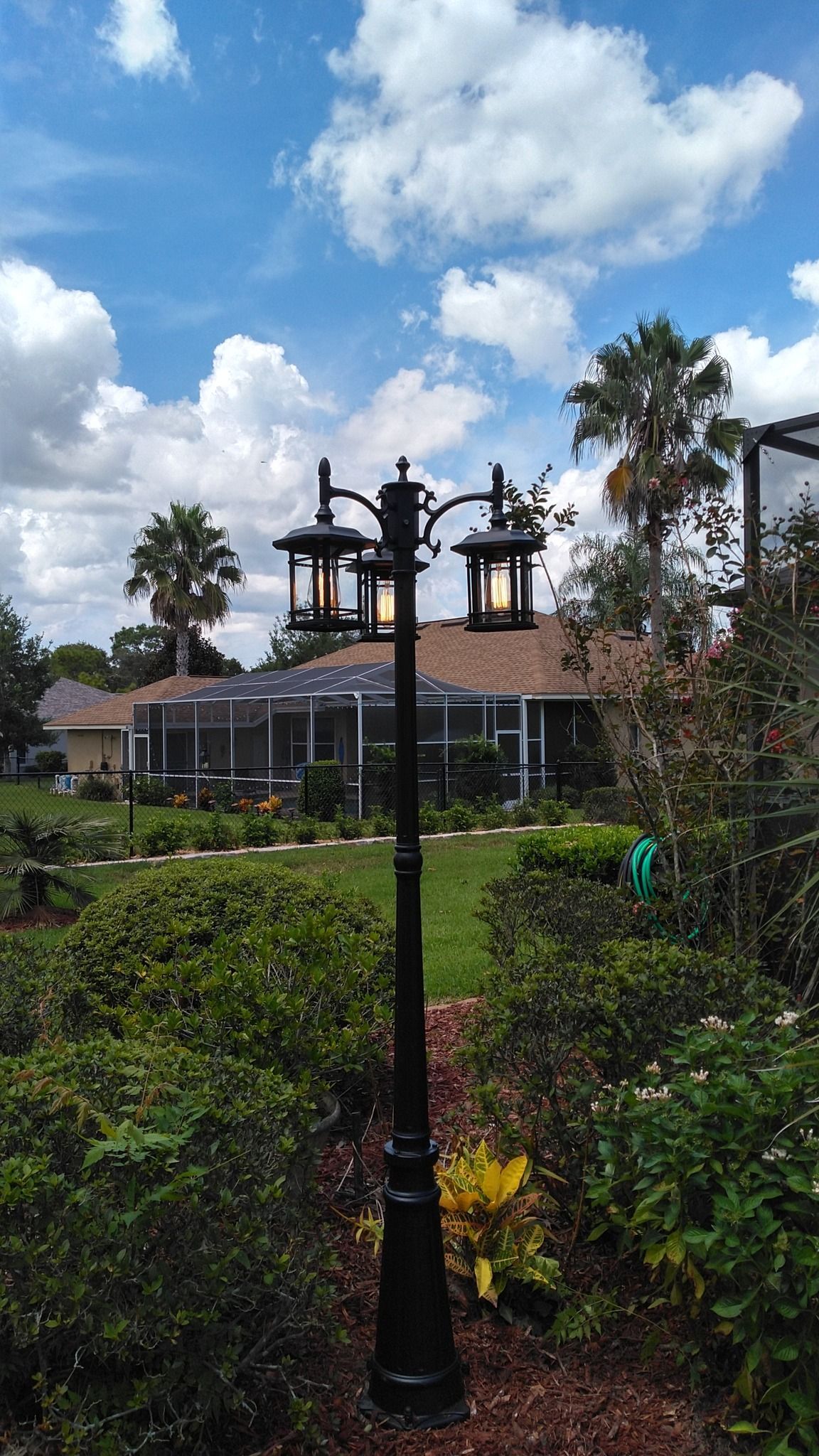 Black lamp post with three lanterns against a backdrop of a blue sky with fluffy clouds, houses, and trees.