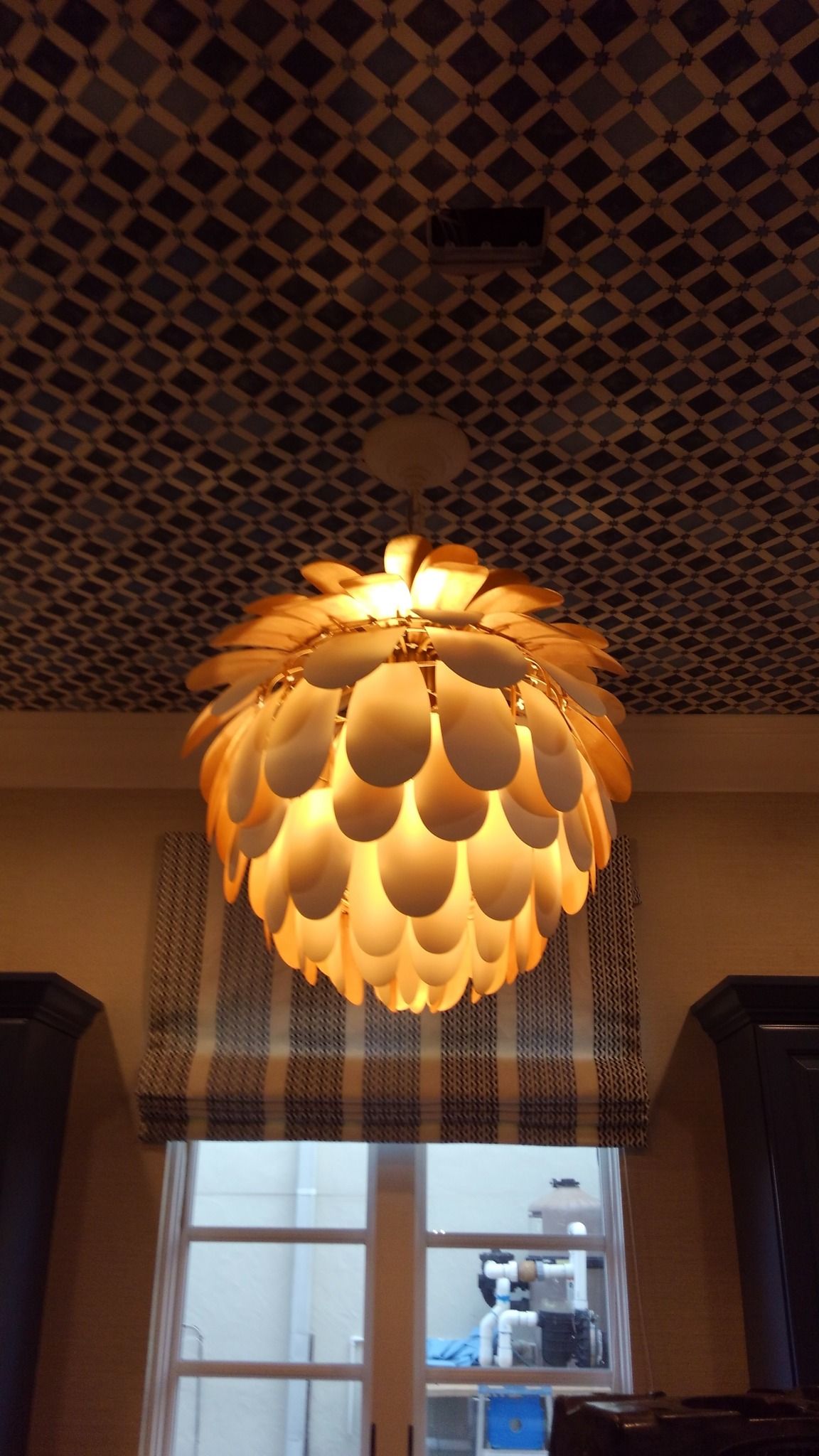 A round, wooden pendant light hangs in front of a window. A patterned ceiling is in the background.