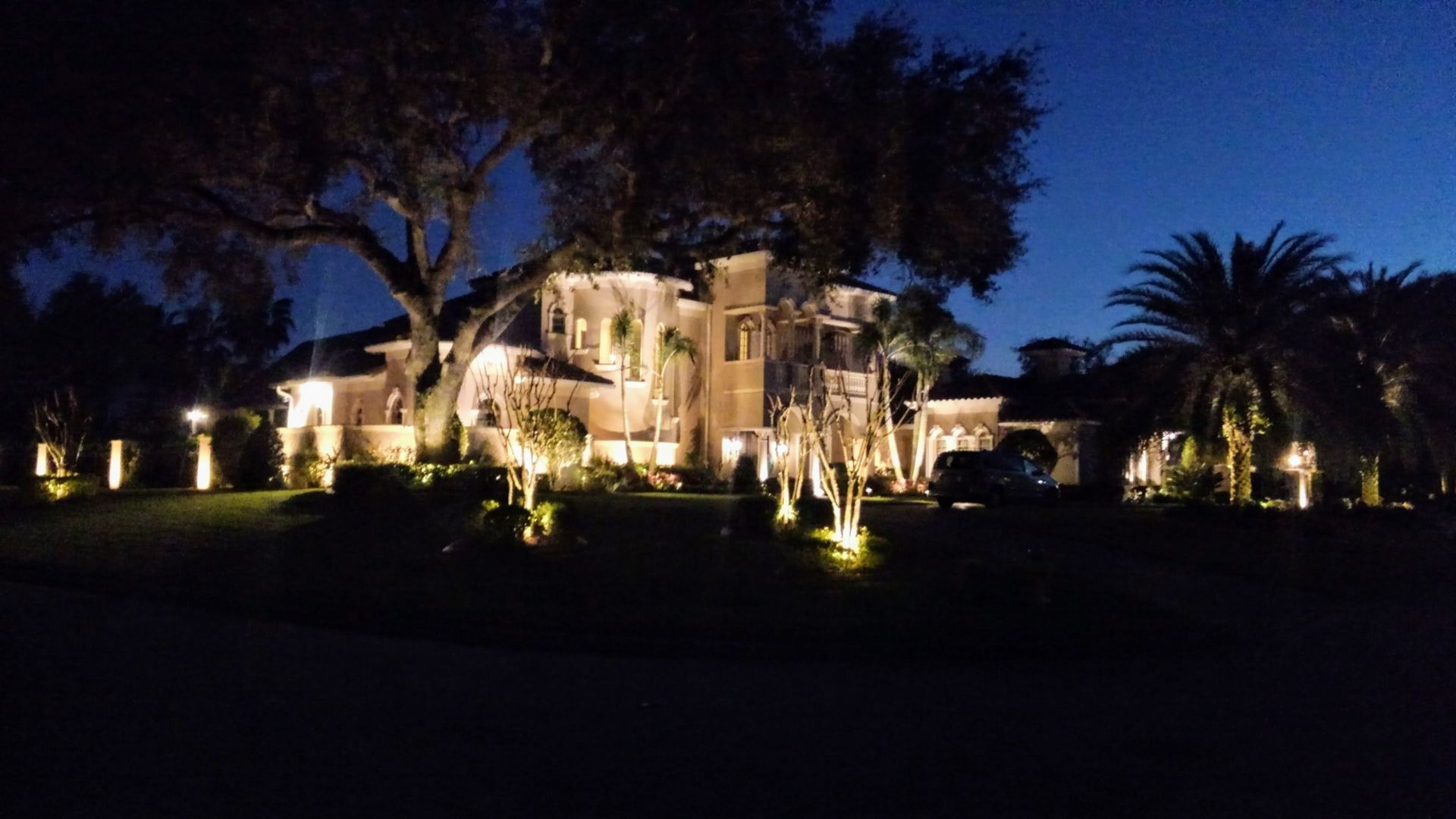 A large, lit-up mansion at night with trees and landscape lighting along a dark street.