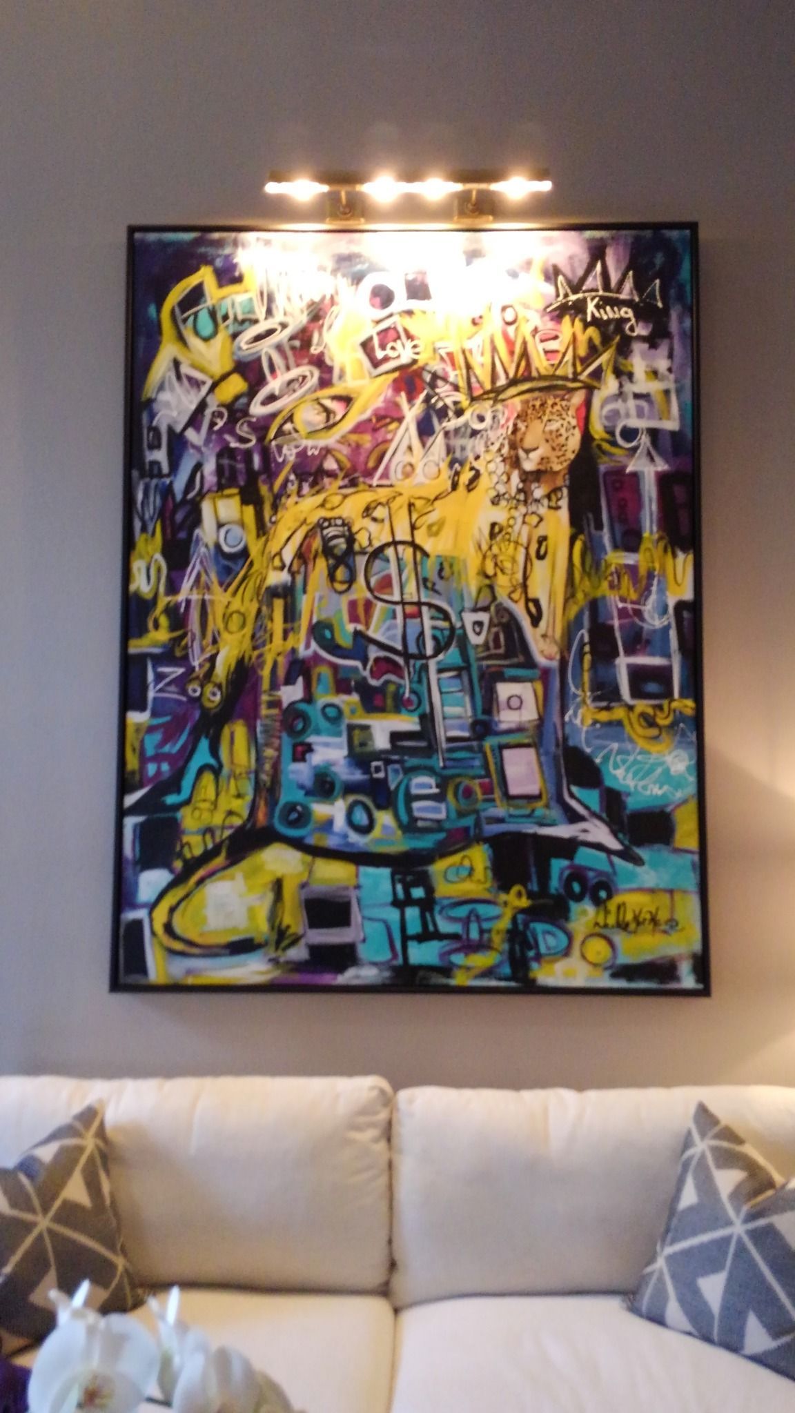 Abstract painting in yellow, blue, and purple hues, above a white sofa, lit by a strip light.