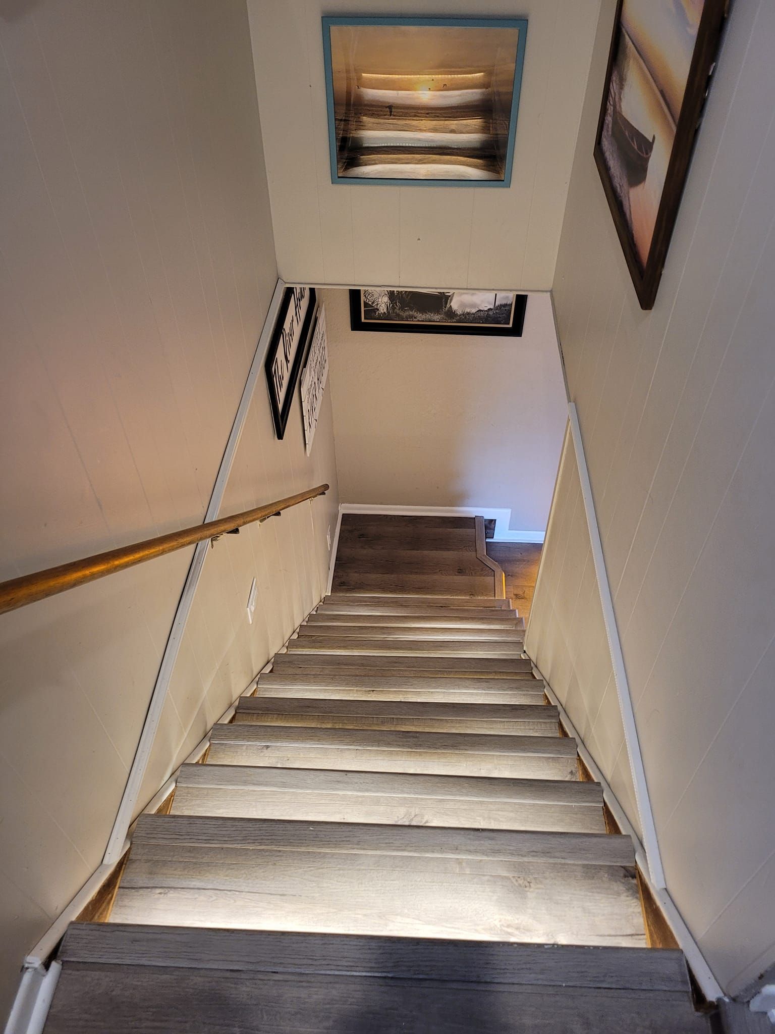 Staircase descending. Wooden steps, white walls, a handrail, and framed pictures.