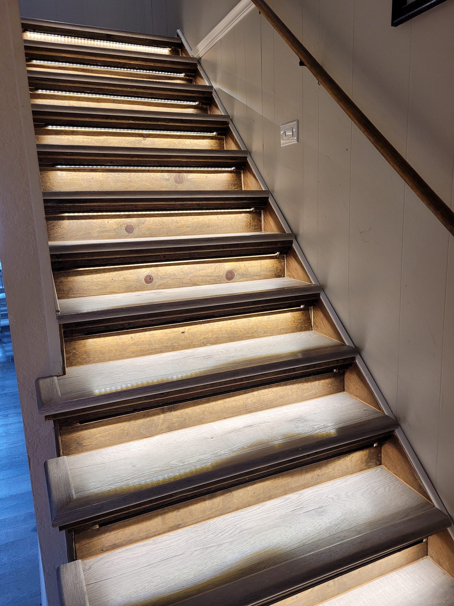 Staircase with built-in step lighting. Wooden steps with warm lights, against a neutral wall and wooden handrail.