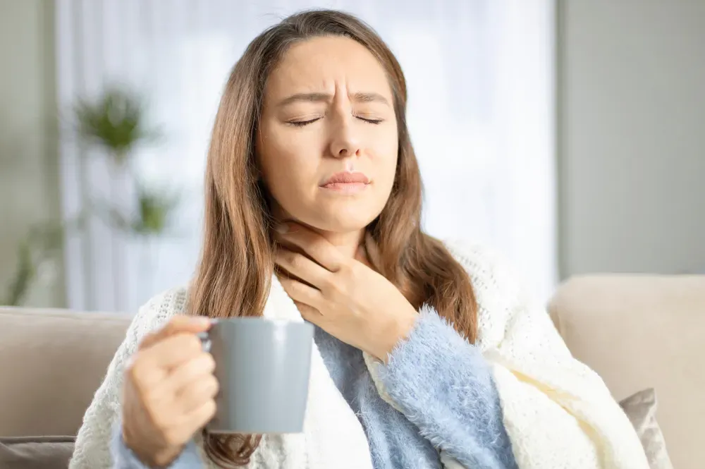 A woman is sitting on a couch holding a cup of coffee and holding her throat.