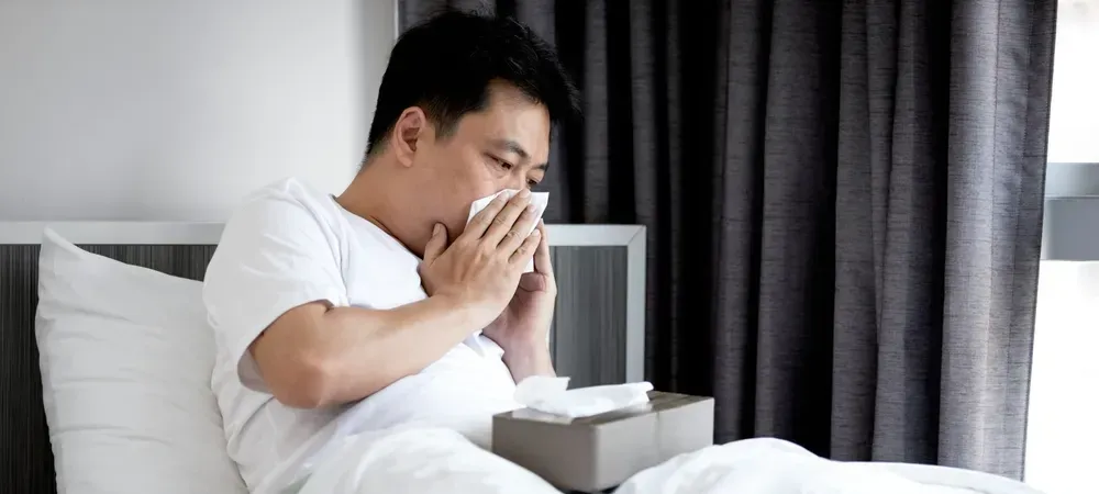 A man is sitting in bed blowing his nose into a napkin.