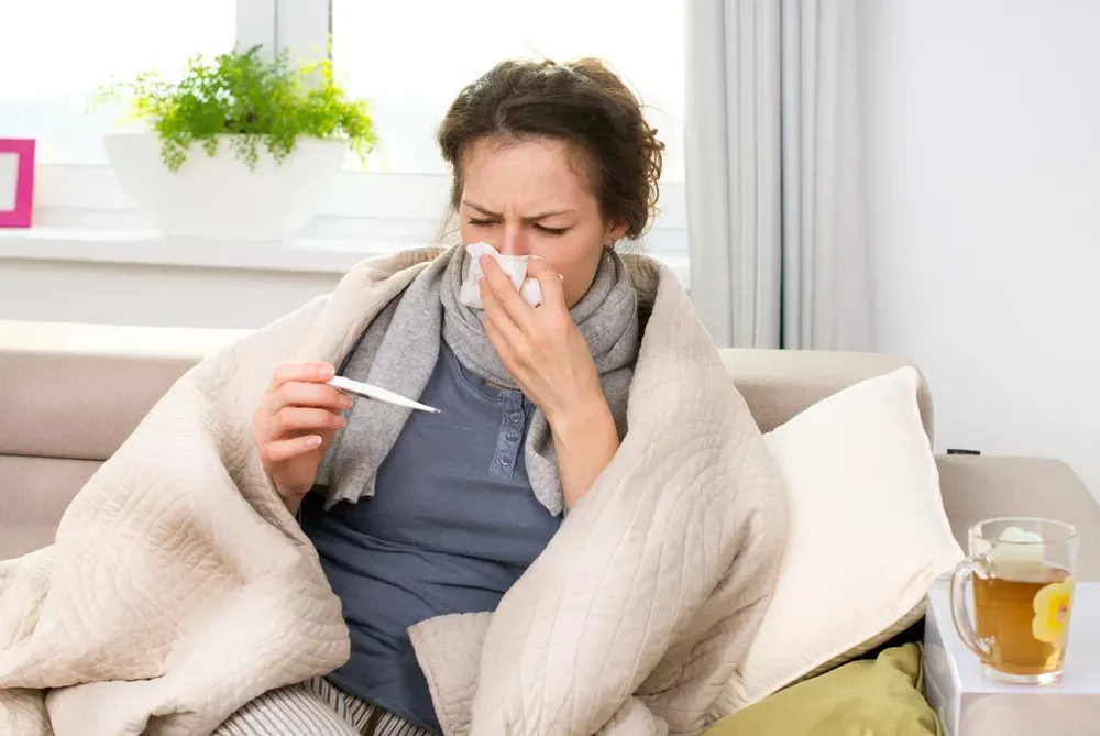 A woman is sitting on a couch with a thermometer in her hand and blowing her nose.