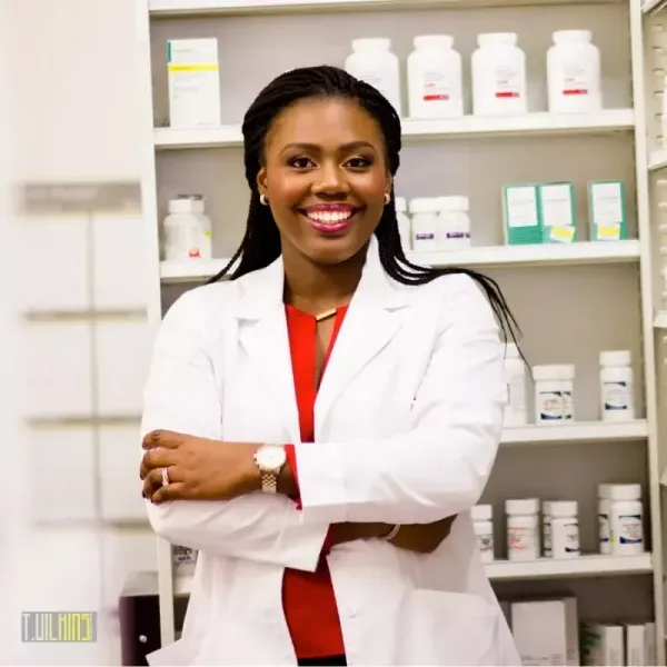 A woman in a lab coat is standing in front of shelves of pills