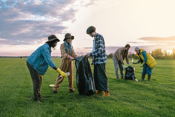 People cleaning up trash in a grassy field; holding black trash bags. Sunny sky in the background.