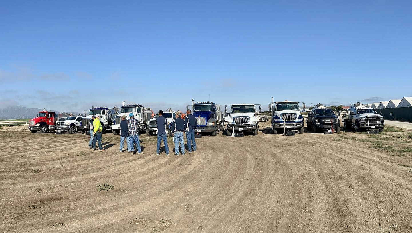 A group of people are standing in front of a row of trucks in a dirt field.