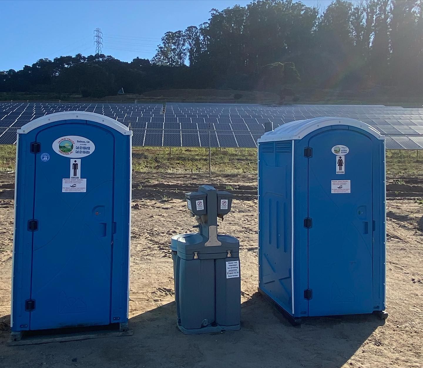 Three blue portable toilets are lined up in front of a field of solar panels