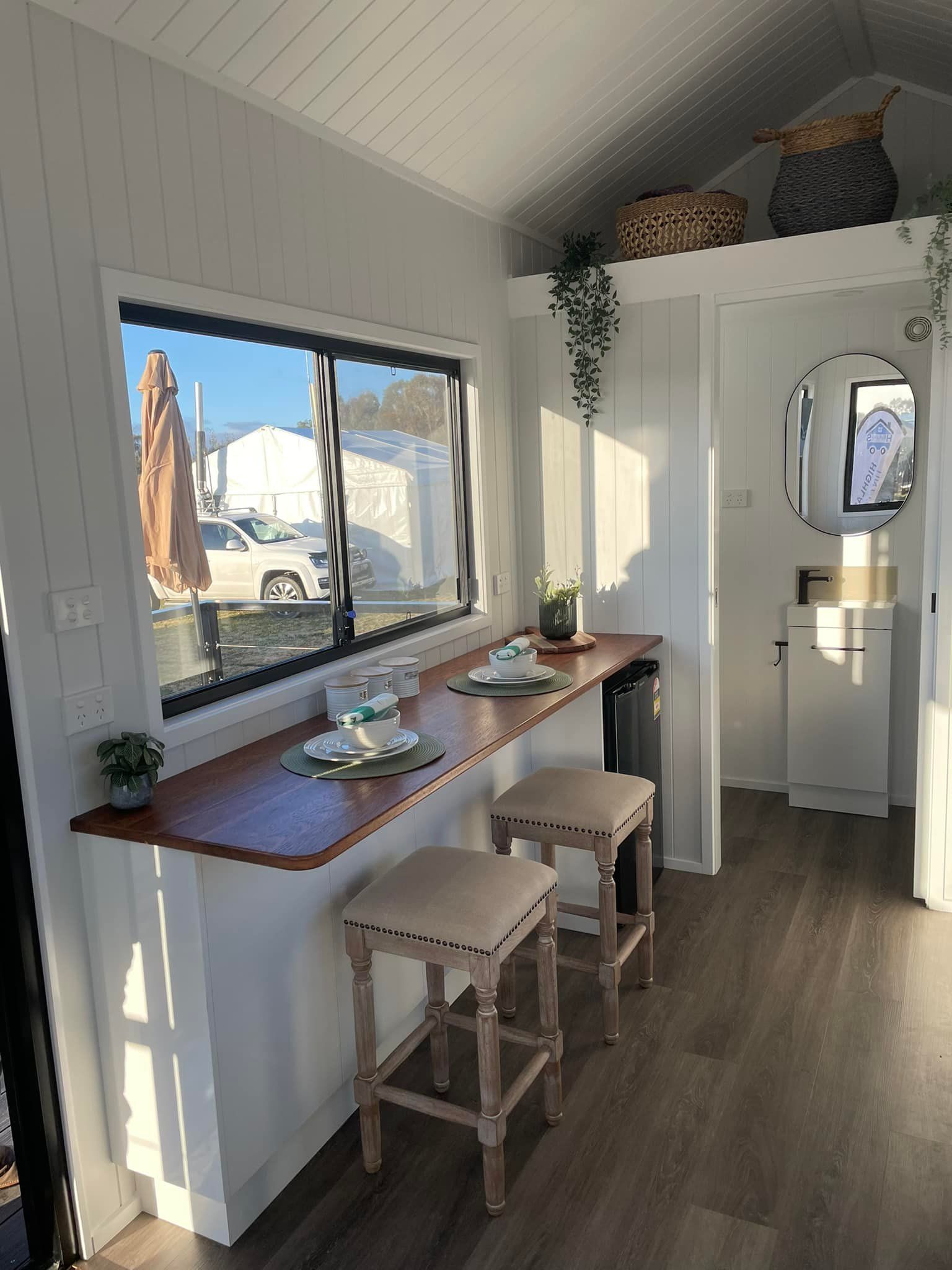Tiny home interior with counter, stools, window overlooking vehicles, white walls, and wood accents.