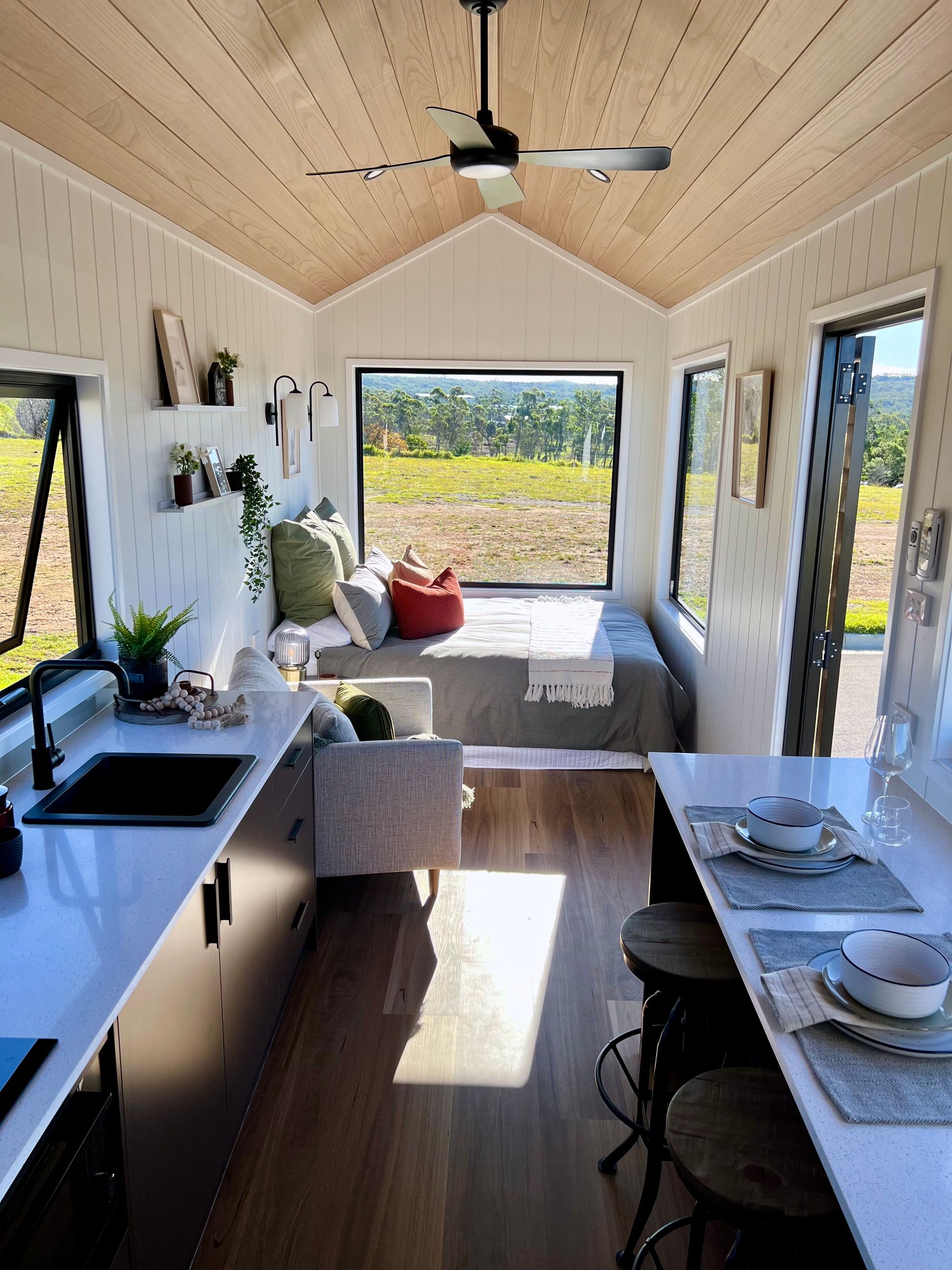 Tiny home interior: kitchen, seating area with large window overlooking a field, wood ceiling and floor.