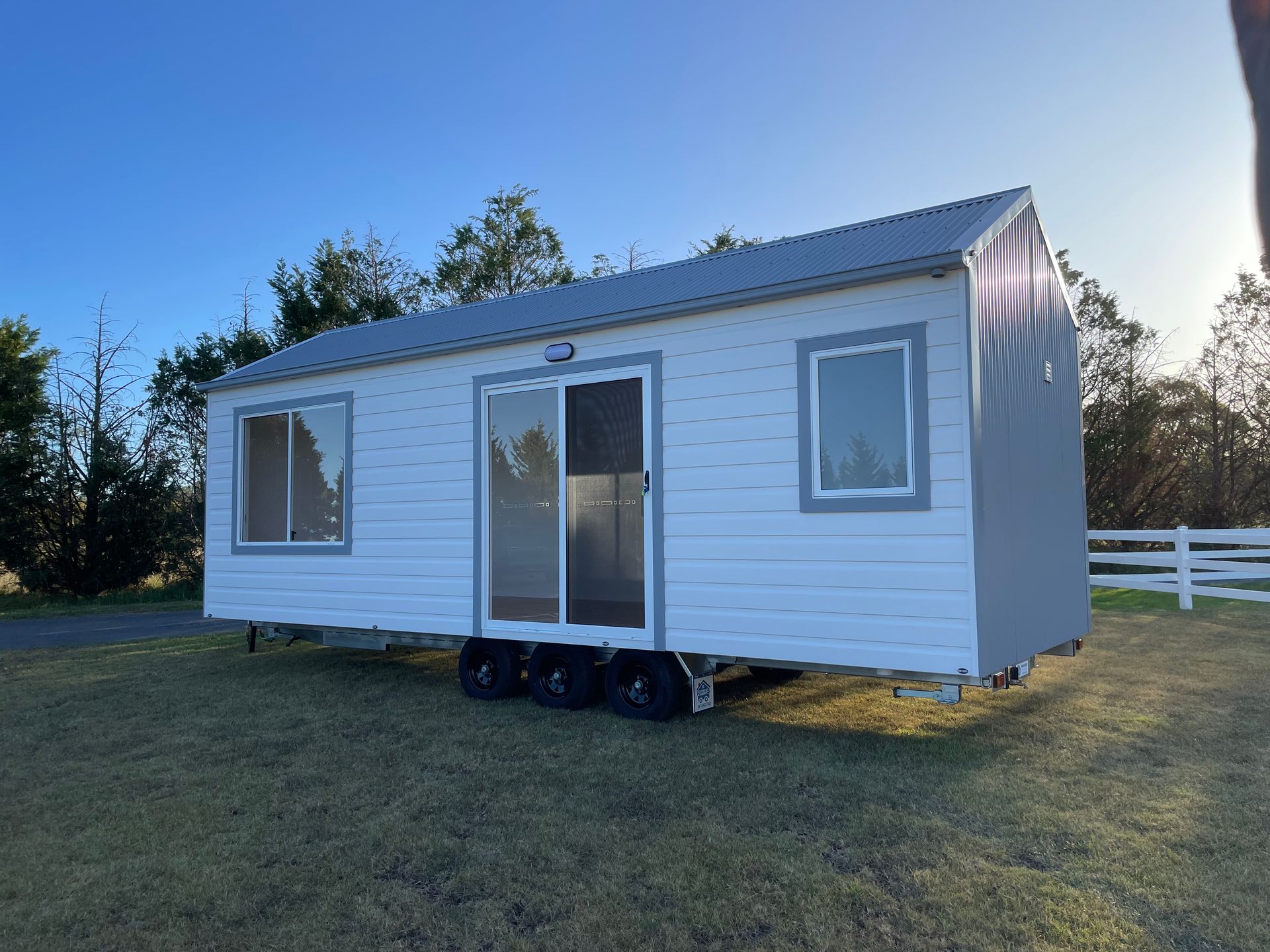 White tiny house on wheels in a grassy field under a blue sky. Sliding glass door, windows.