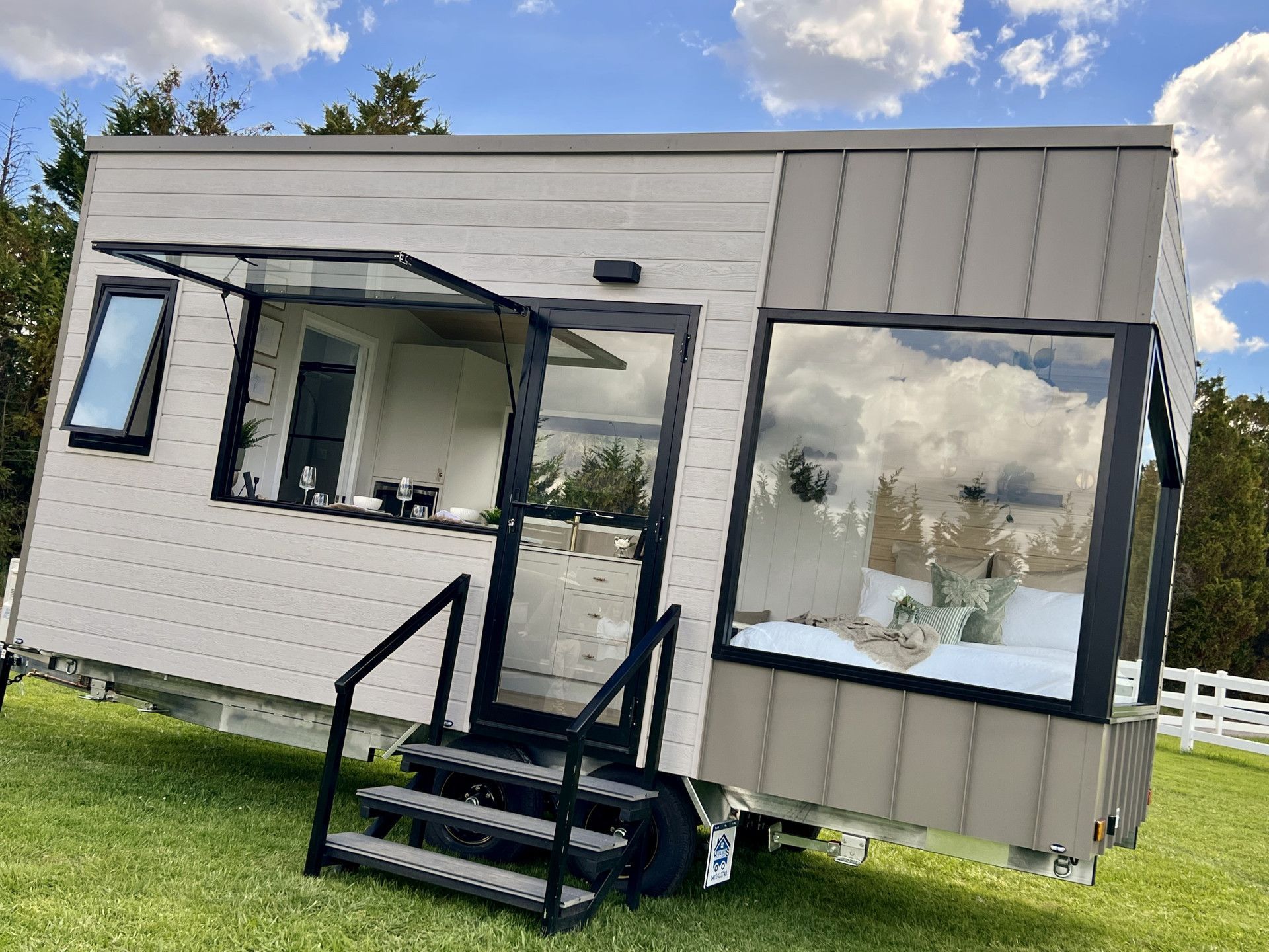 Tiny house on wheels with large windows and a black door, set on green grass under a blue sky.