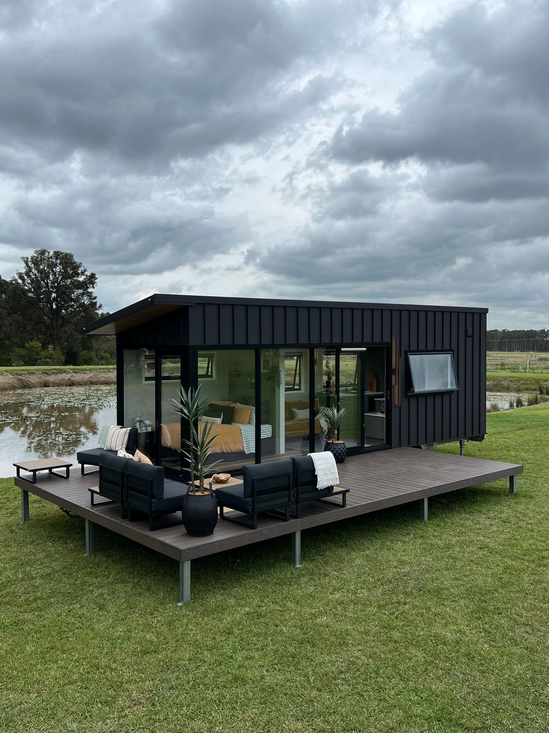 Modern black cabin with large windows and a deck overlooking a grassy area and pond under a cloudy sky.