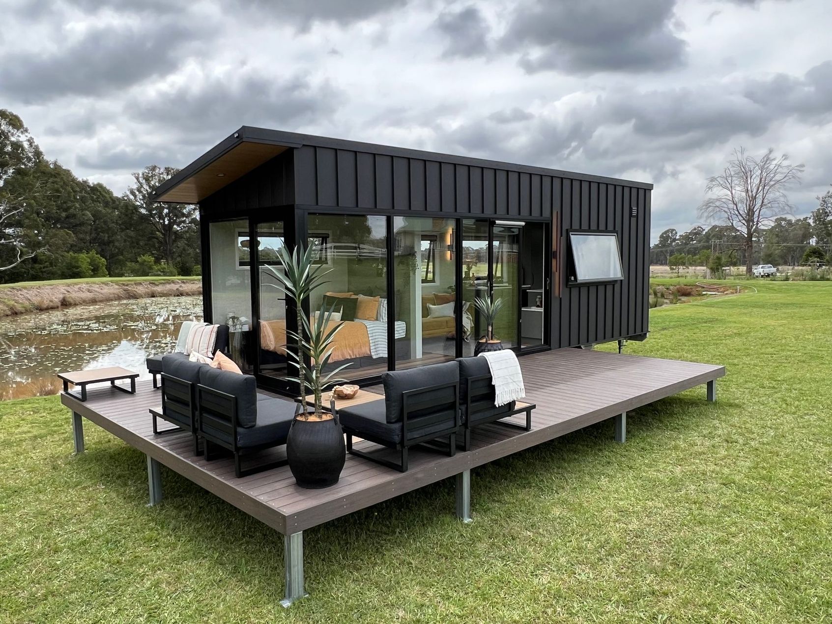 Modern black cabin with glass walls and deck, on green grass, next to a small pond.