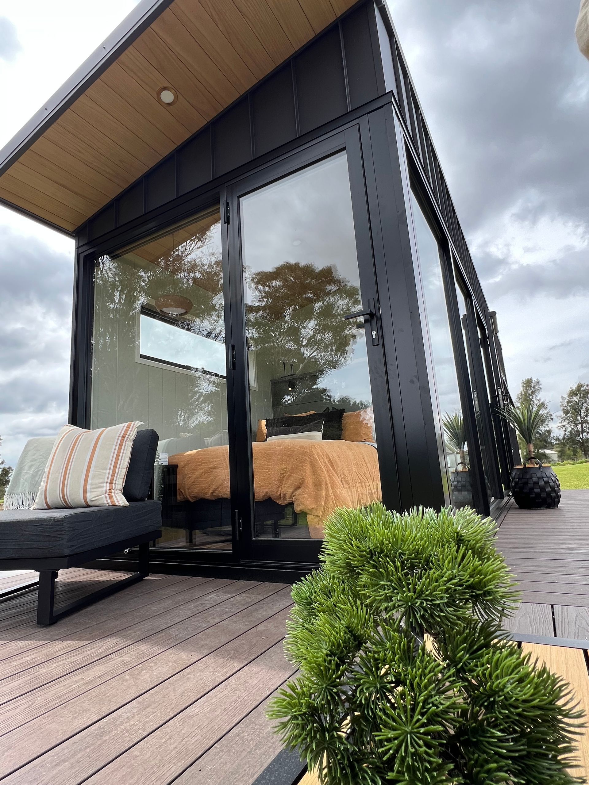 Modern cabin with glass walls and dark trim, wooden deck, outdoor seating, and lush greenery.
