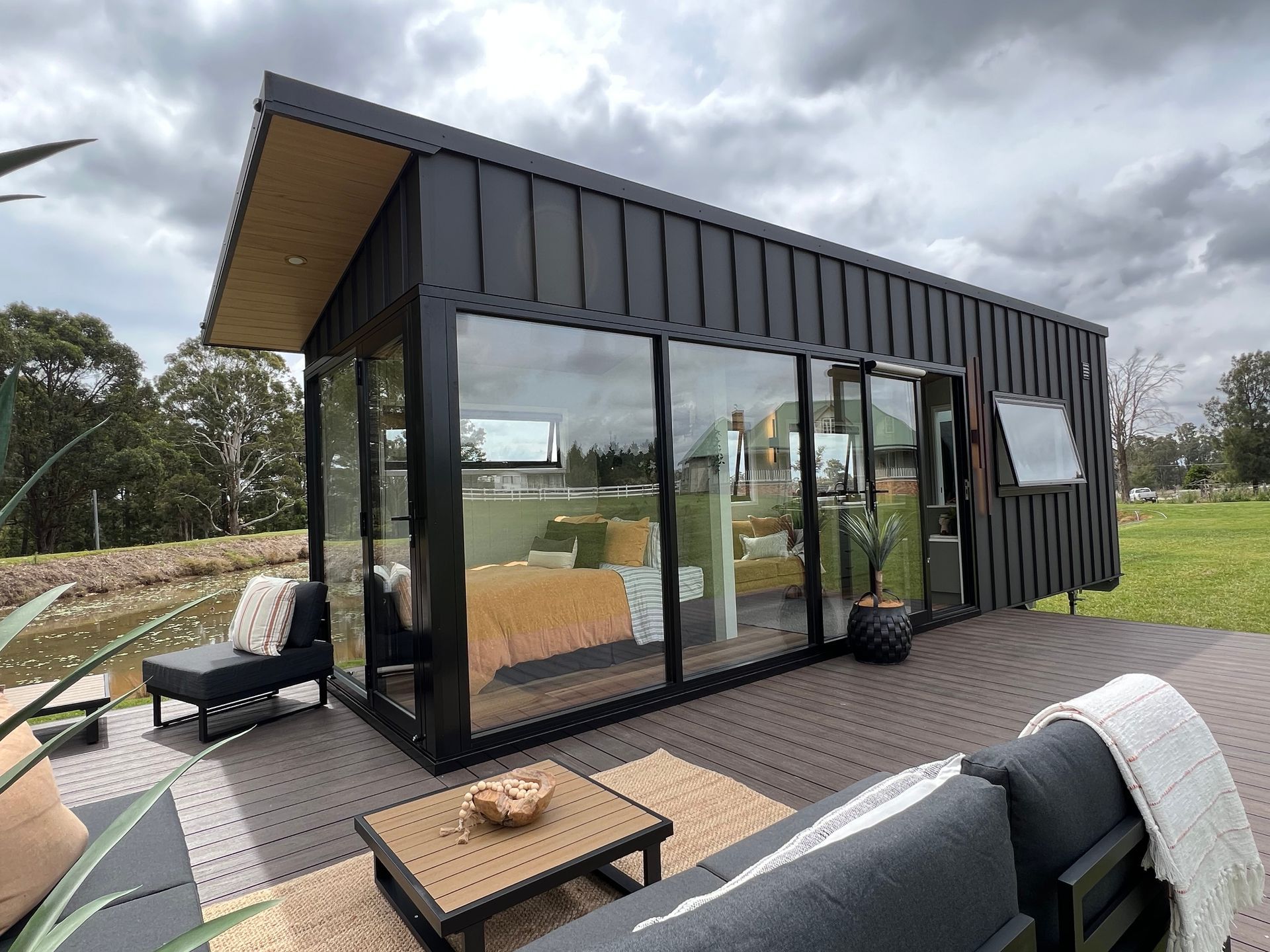Modern tiny home with black exterior, glass walls, and outdoor seating on a deck overlooking a pond.