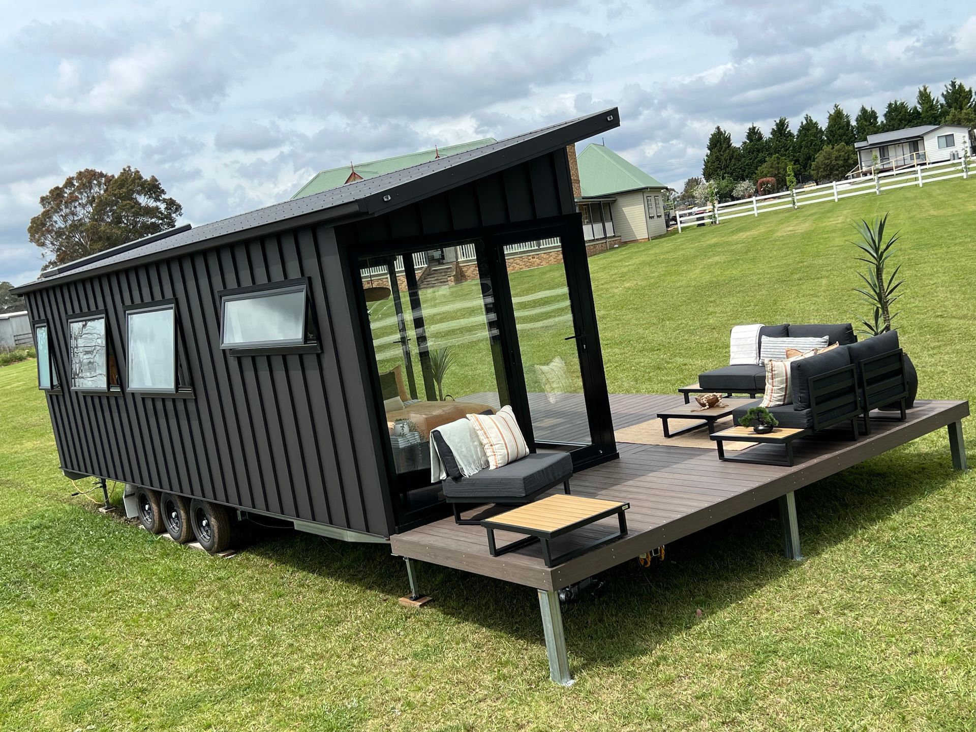 Black tiny home on wheels with a deck and outdoor seating on a grassy hill.