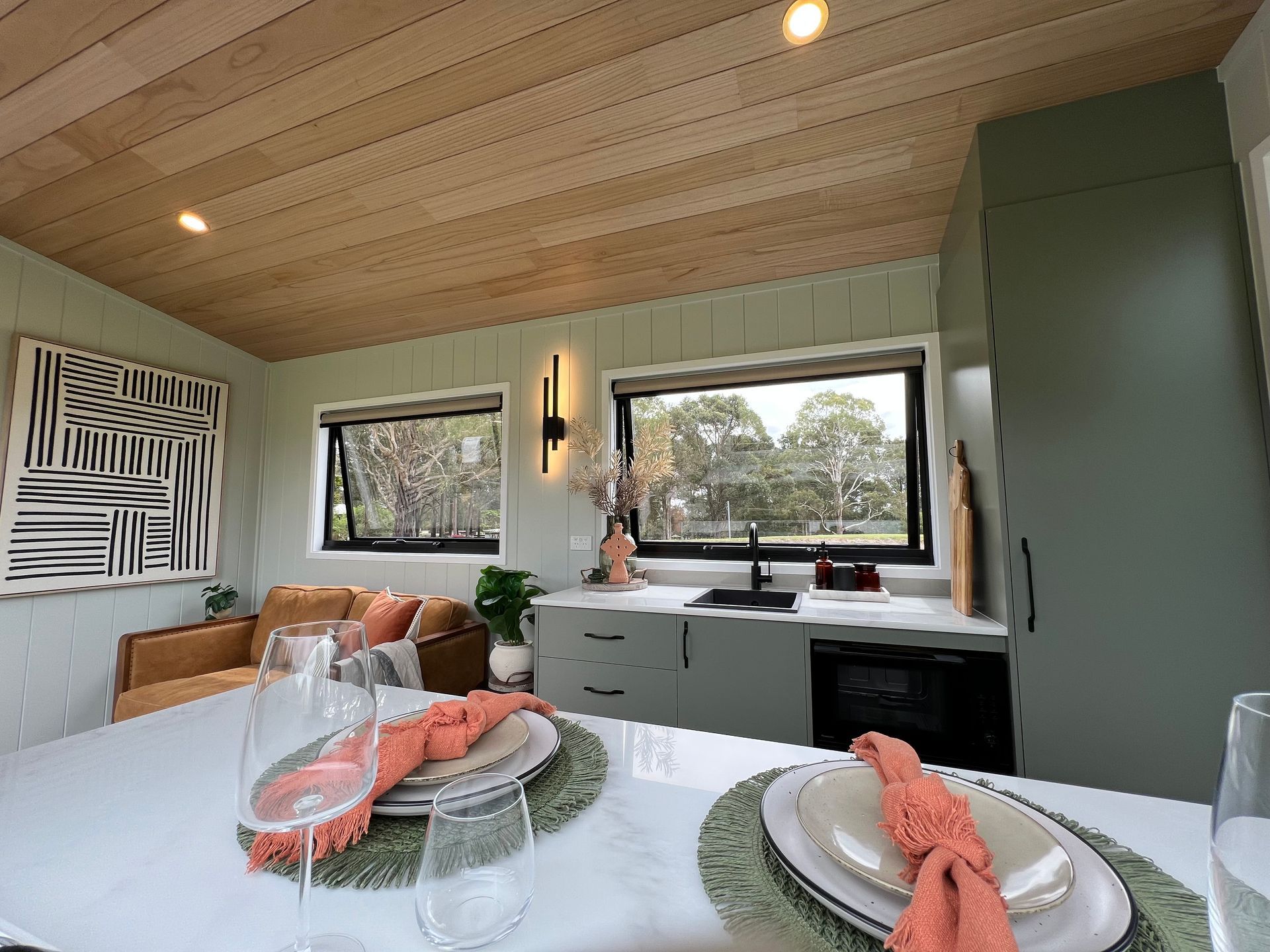 Interior of tiny home kitchen/dining area. Green cabinetry, white countertops, black accents, and art on wall.