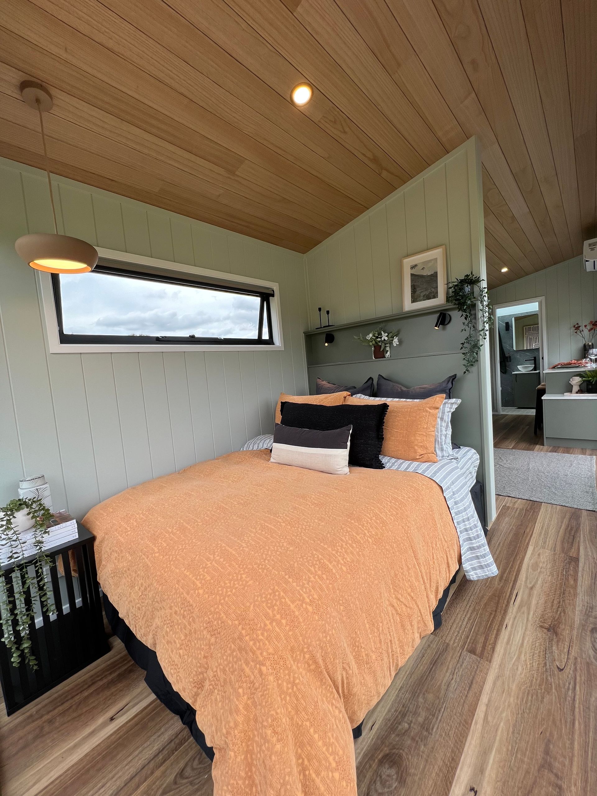 Cozy bedroom with twin bed, green/red pillows, large window overlooking a field, white walls, and black sconces.