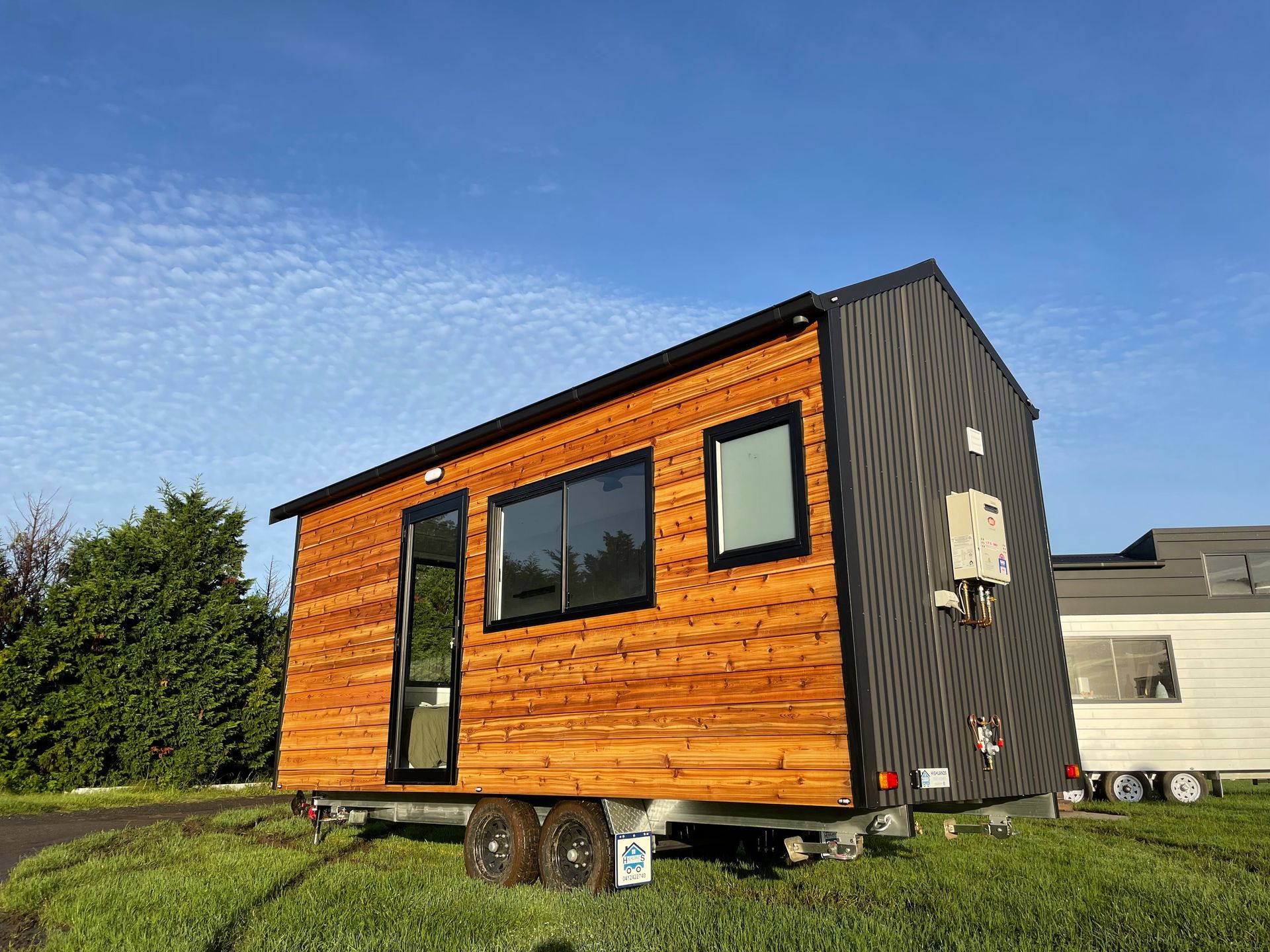 Tiny wooden house on wheels, with black trim, set on a green lawn under a blue sky.