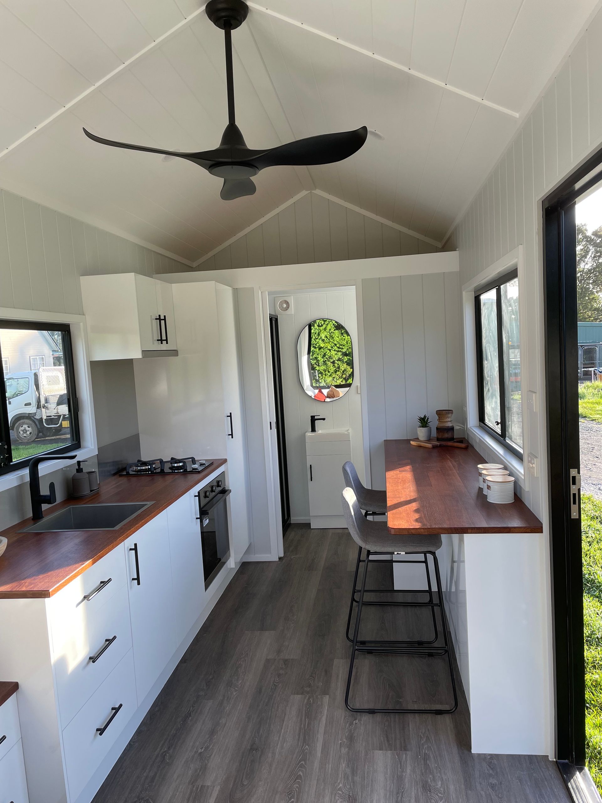Interior of a tiny house. White walls, kitchen with wooden countertop, a bar, and a ceiling fan.