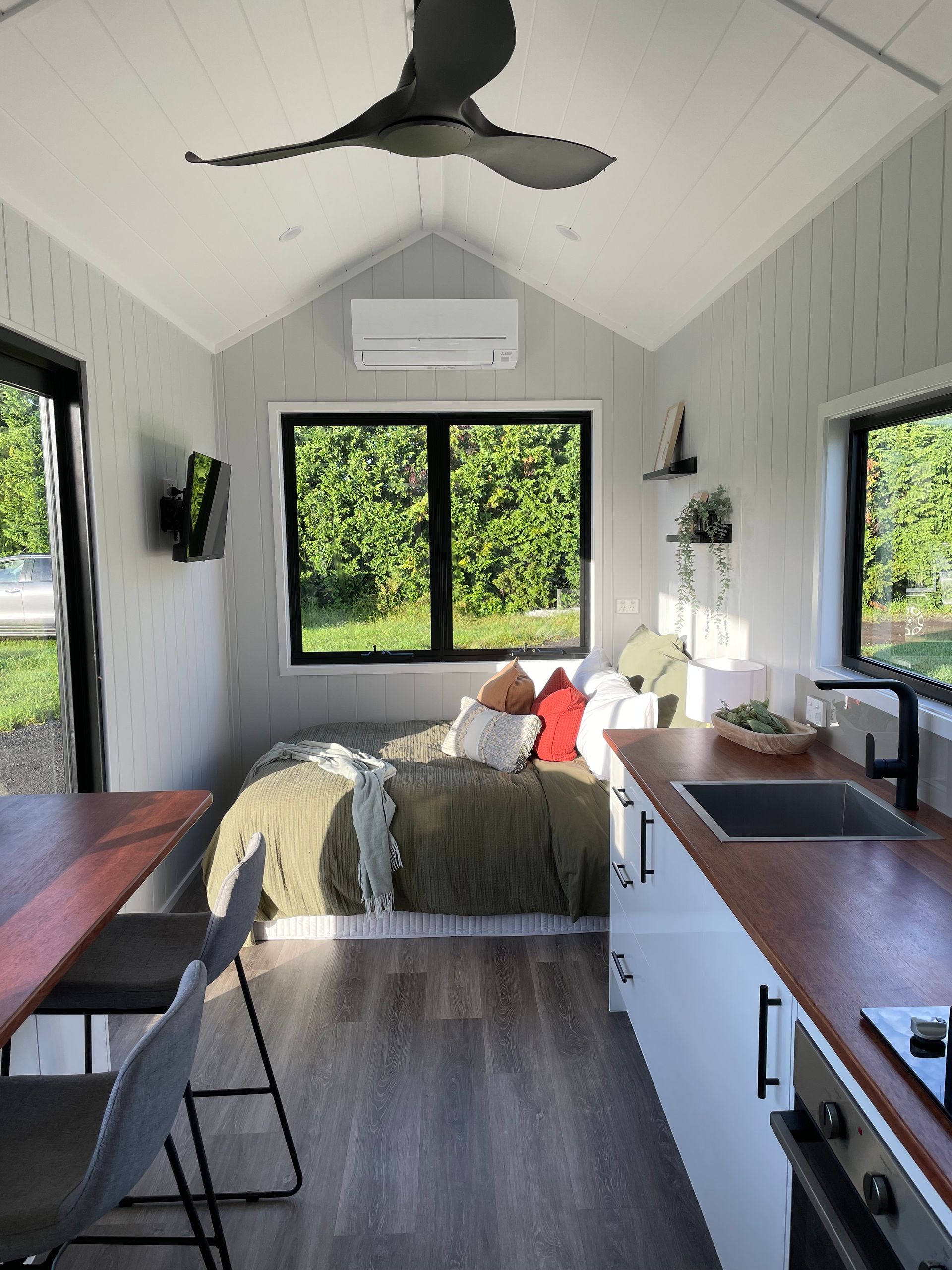 Tiny home interior with bed, kitchen, and small dining area; windows overlook lush green foliage.