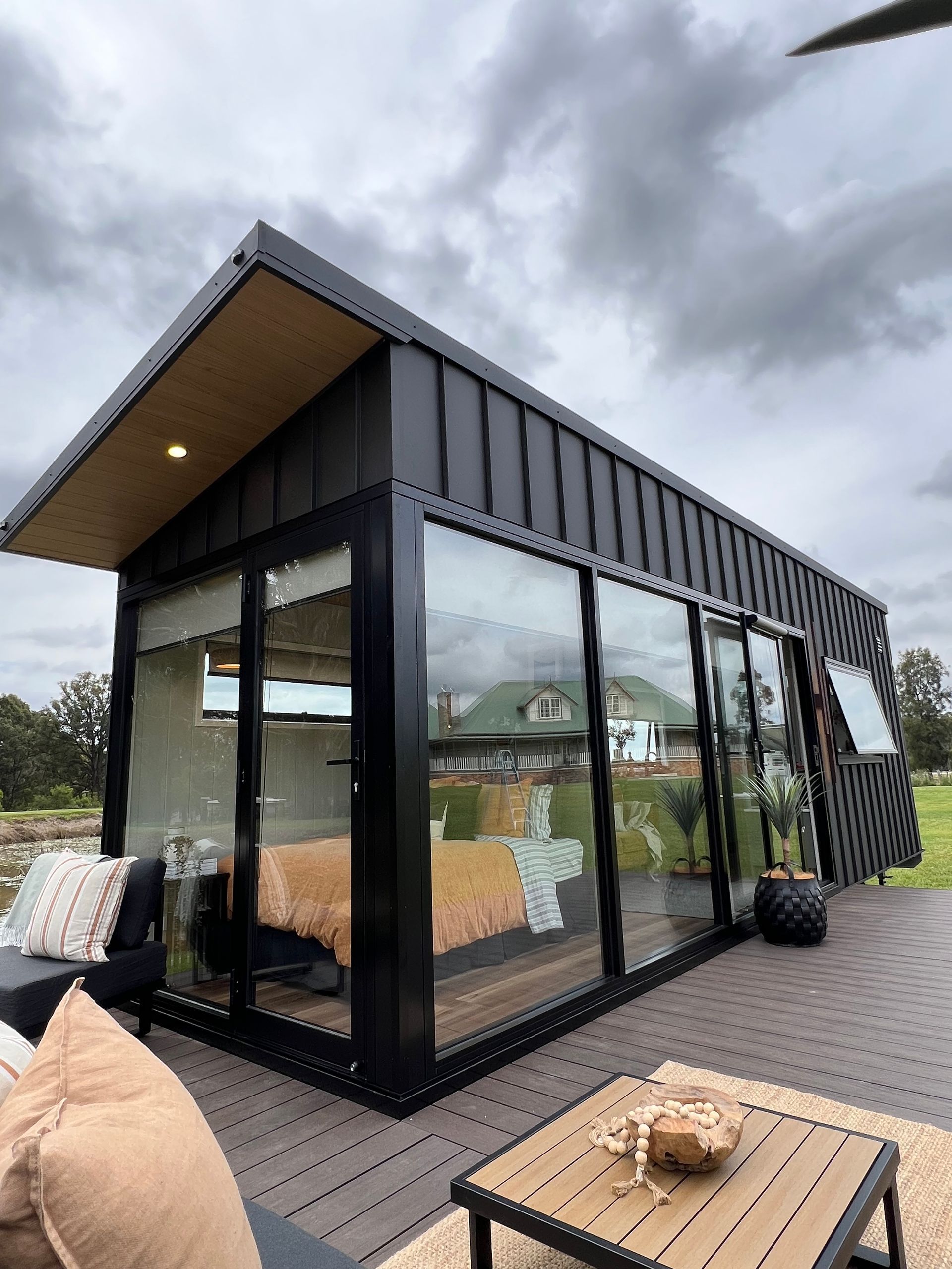Modern black cabin with glass walls, outdoor seating, and a cloudy sky.