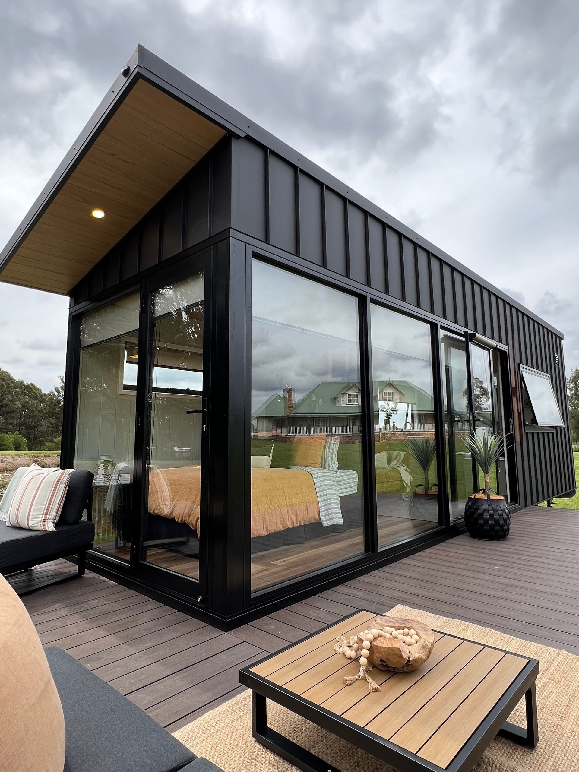 Modern black tiny home with glass walls, outdoor deck, and cloudy sky.