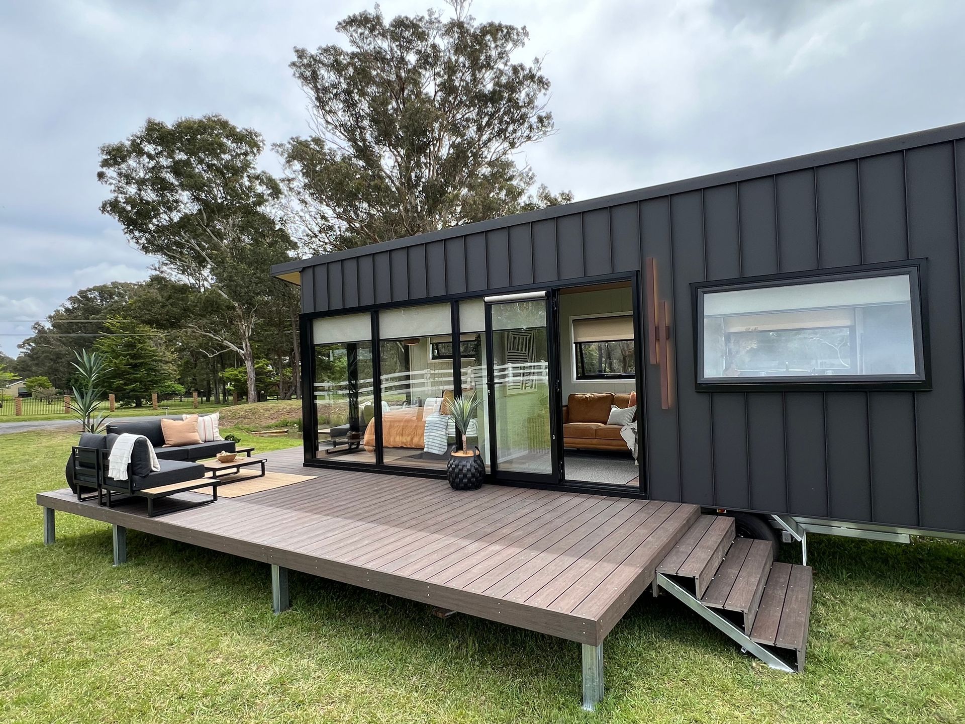 Dark tiny house with a deck on a grassy area, sliding glass doors, and a small window.