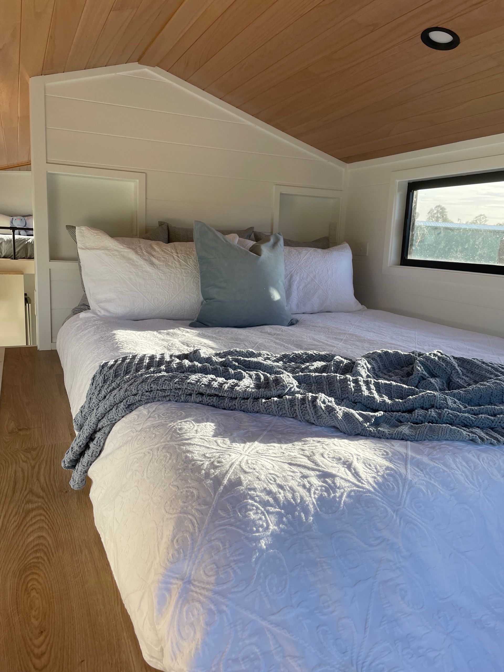 Bedroom with white bed, blue blanket and pillow, natural wood ceiling, and black window.