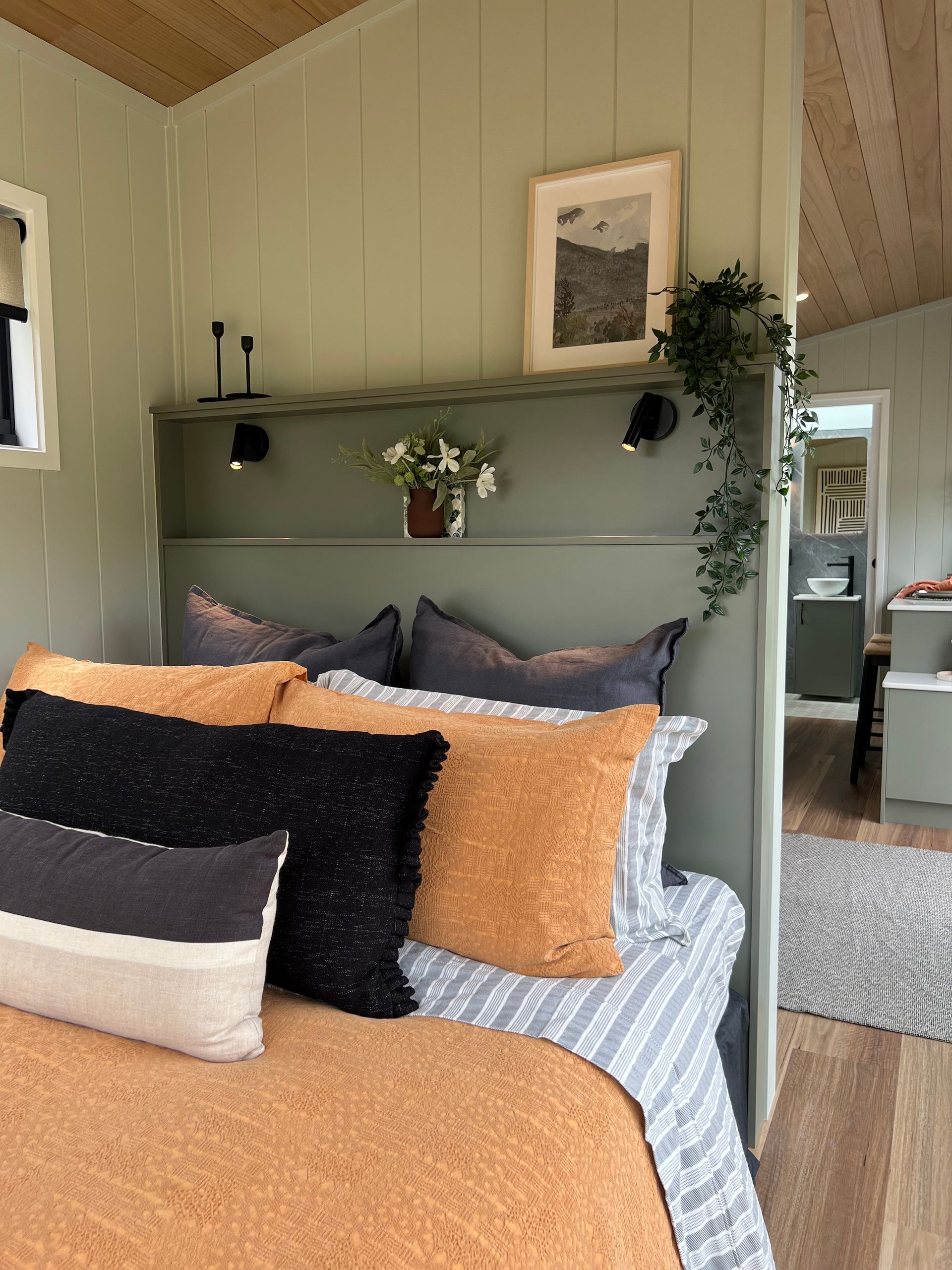 Cozy bedroom with sage green walls, orange bedding, and decorative pillows. Headboard with shelf, artwork, and hanging plant.
