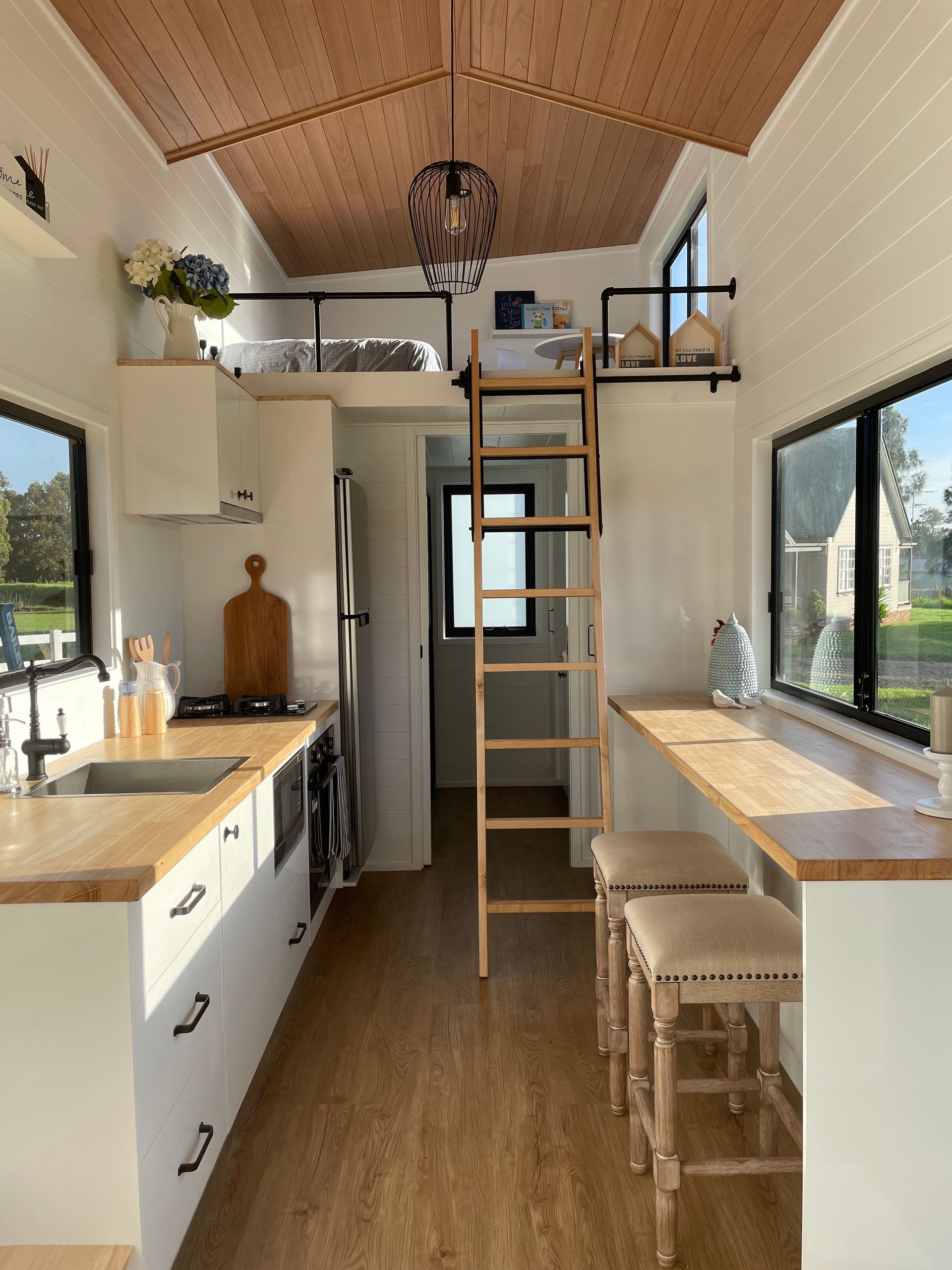 Interior of a tiny house with a kitchen, dining area, and loft. Features light wood tones and white walls.