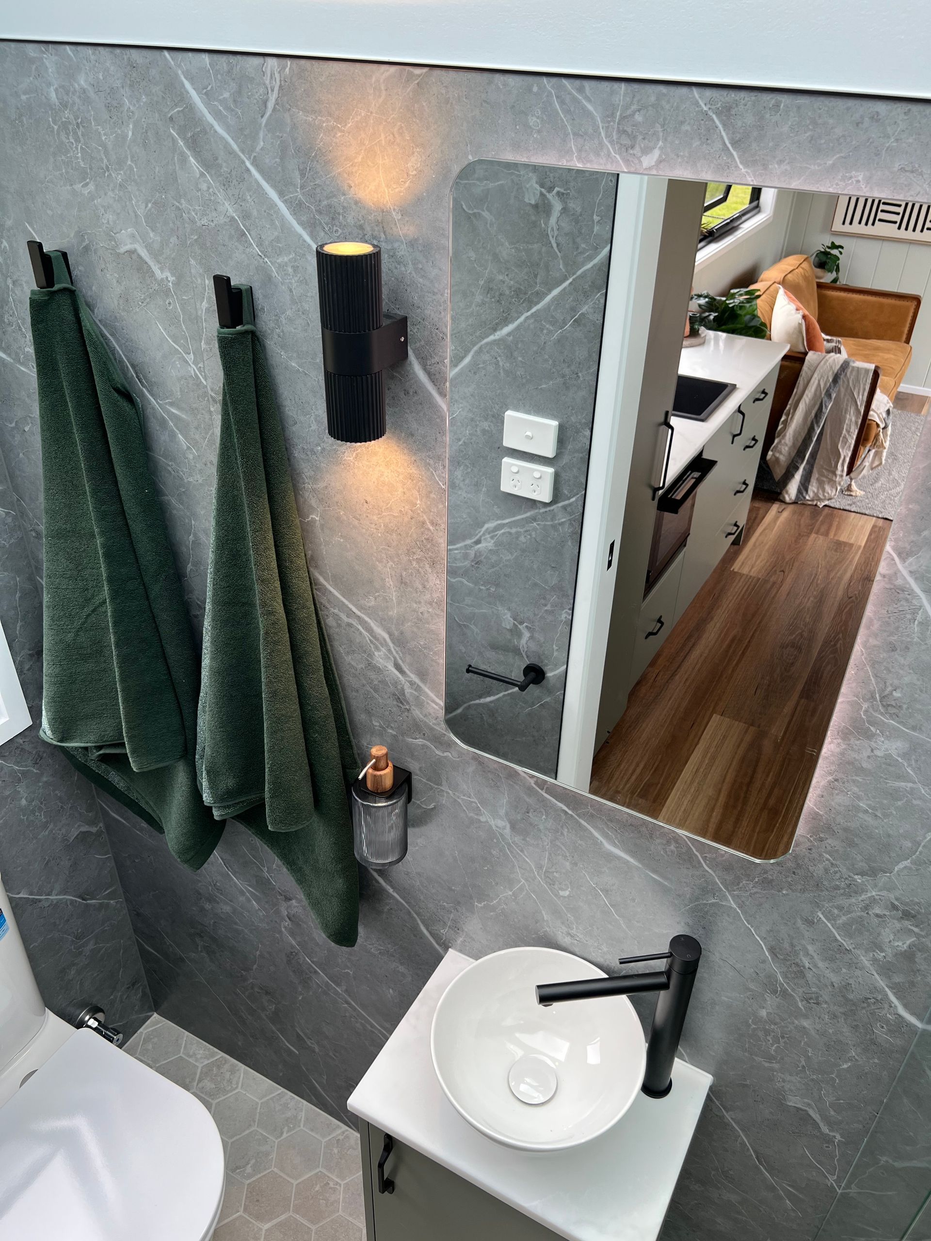 Modern bathroom with gray tiled wall, green towels, black fixtures, and a mirror reflecting a kitchen.