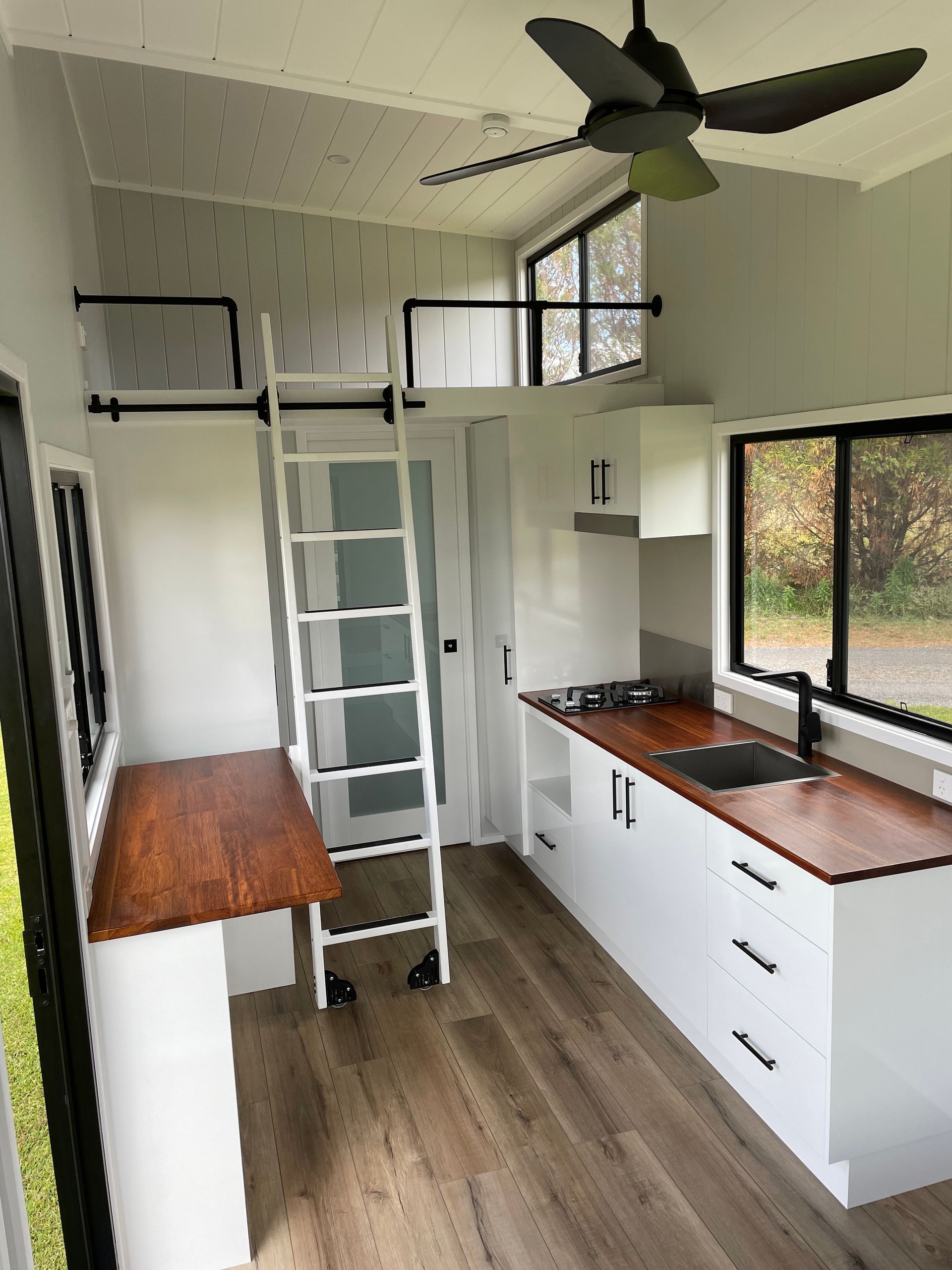 Interior of a tiny house with a loft, kitchen, desk, and ladder. White and brown color scheme.