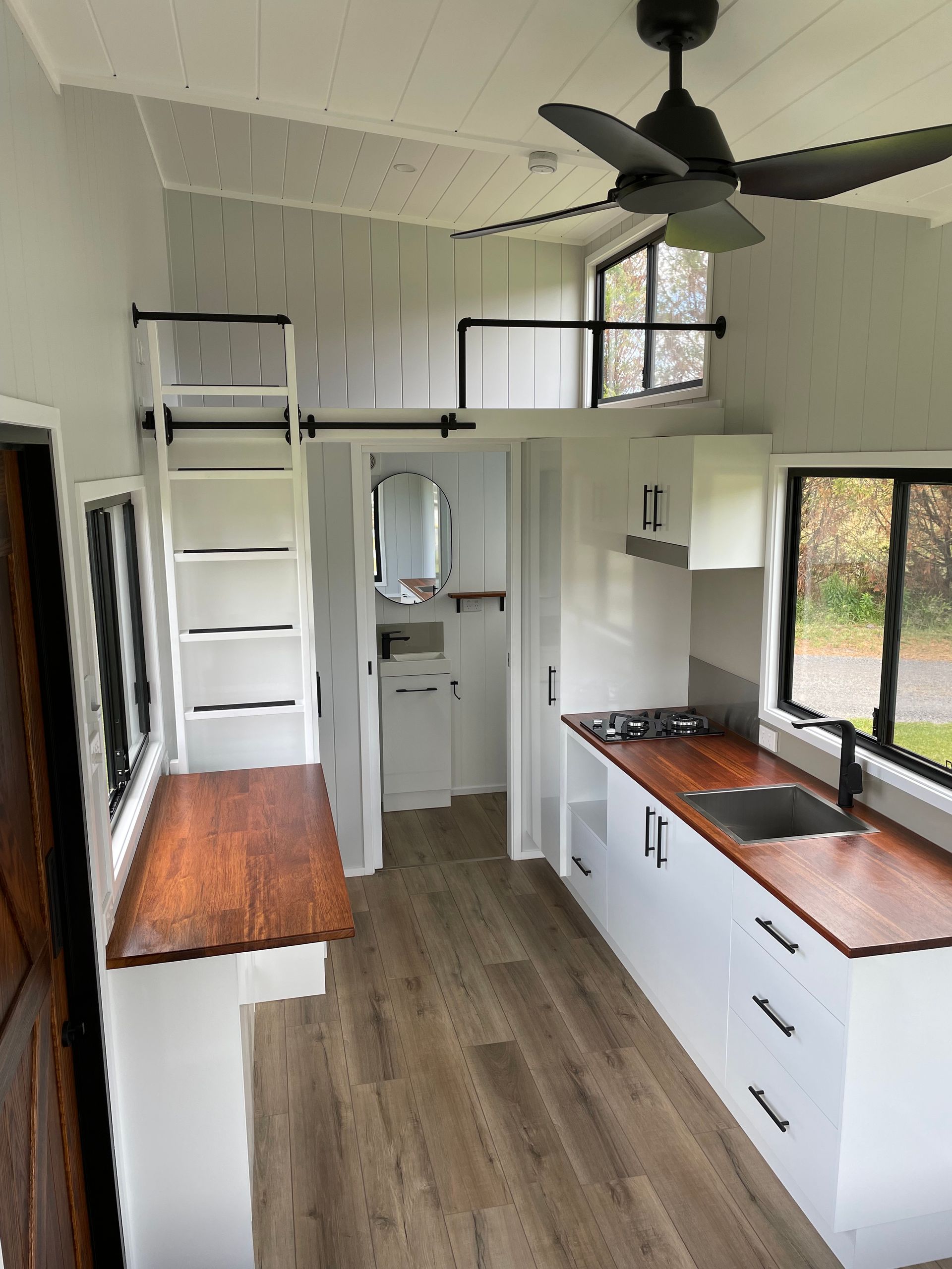 Interior view of a tiny home with a kitchen, loft, and a small bathroom. White walls, wood countertops, and dark trim.