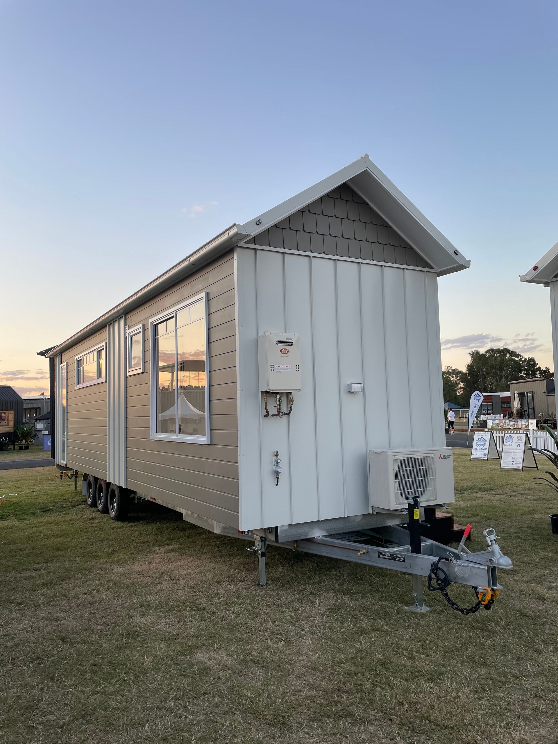 Tiny home on trailer, beige and white exterior, parked on grass at dusk.