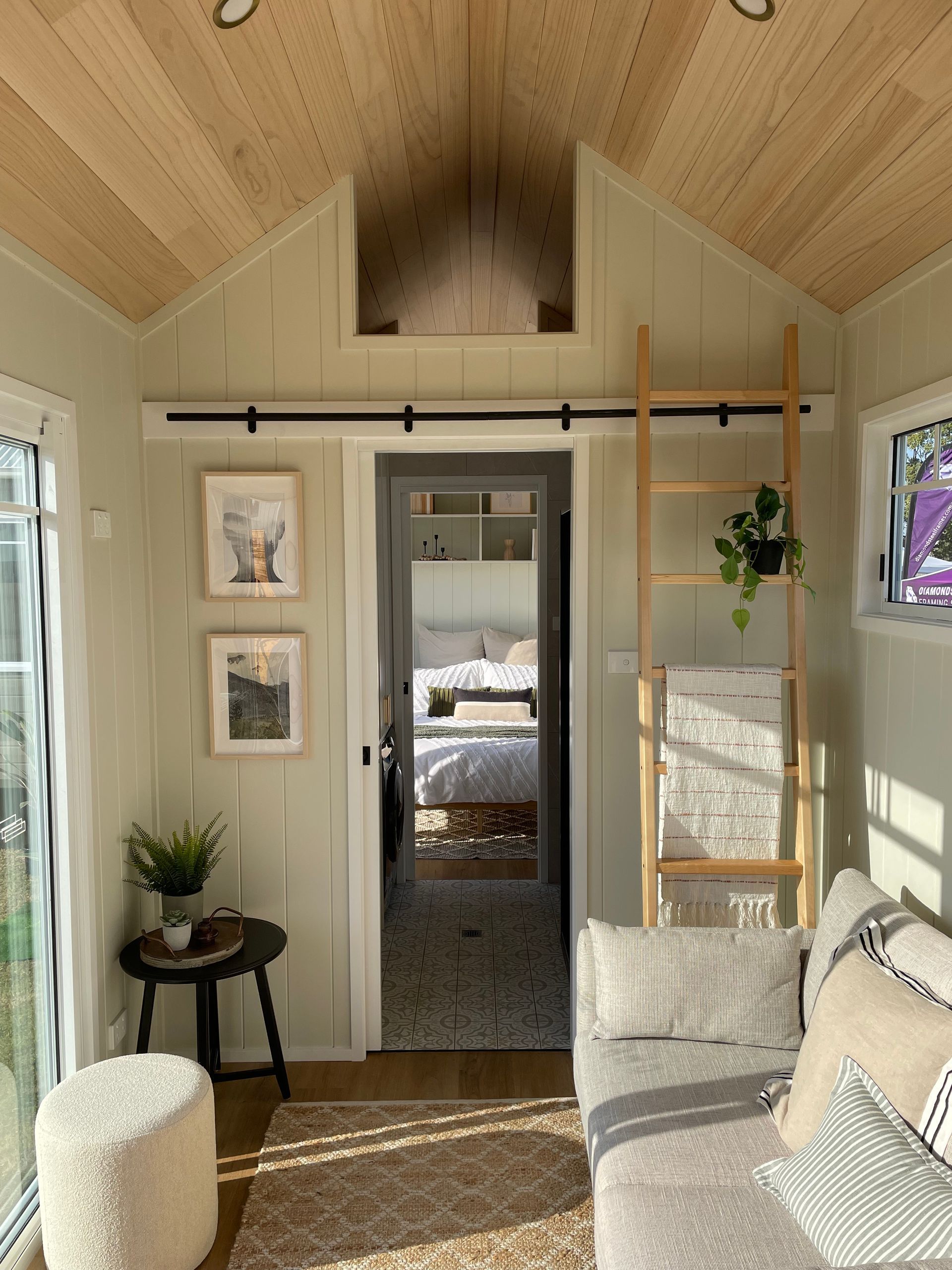 Cozy, light-filled tiny home interior with sofa, ladder, art, and doorway to a bedroom.