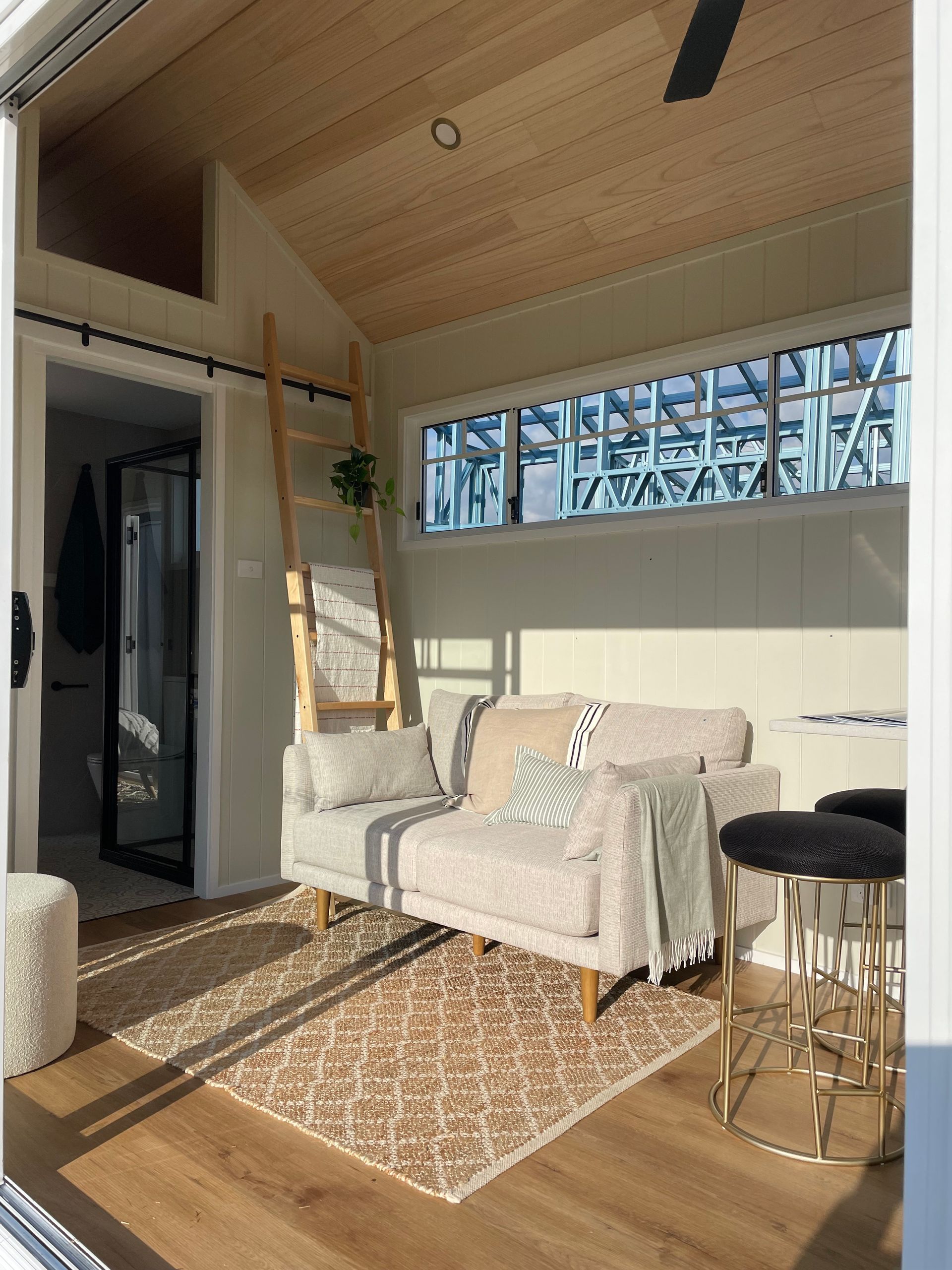 Cozy sunlit tiny home interior: sofa, patterned rug, ladder, open door to bathroom, wood ceiling, and flooring.