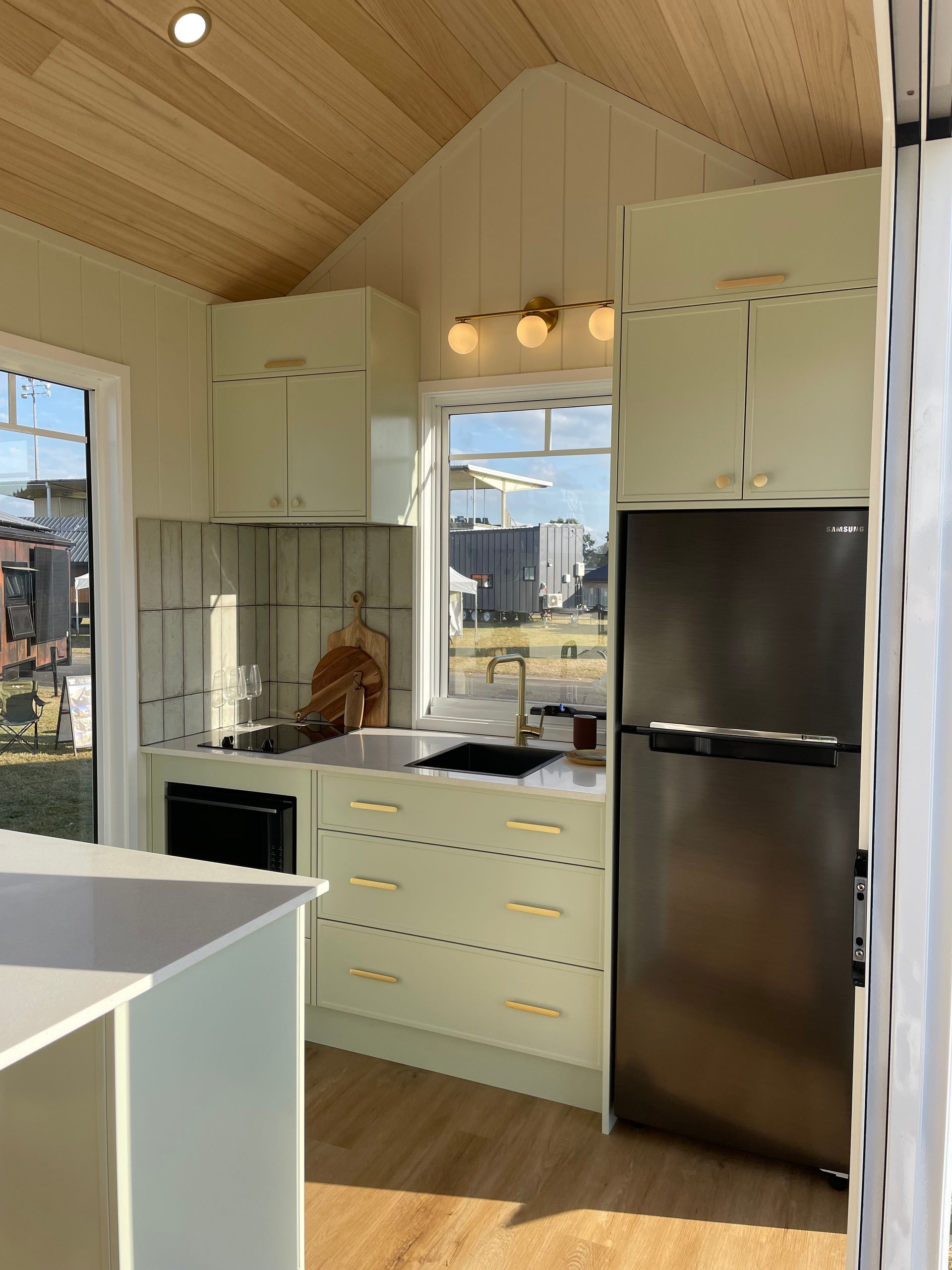 Cozy tiny kitchen with sage green cabinets, gold hardware, and a black refrigerator. Sunlight streams in from the window.
