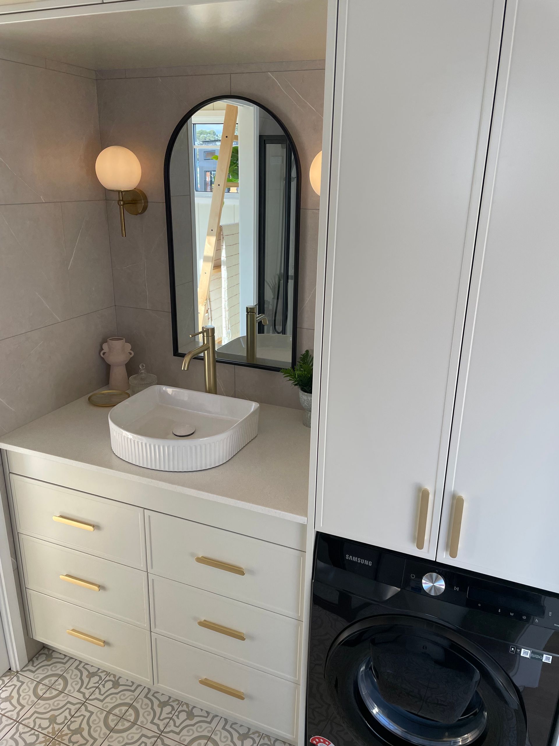 Bathroom with white cabinets, gold hardware, black washer, arched mirror, and sconces.