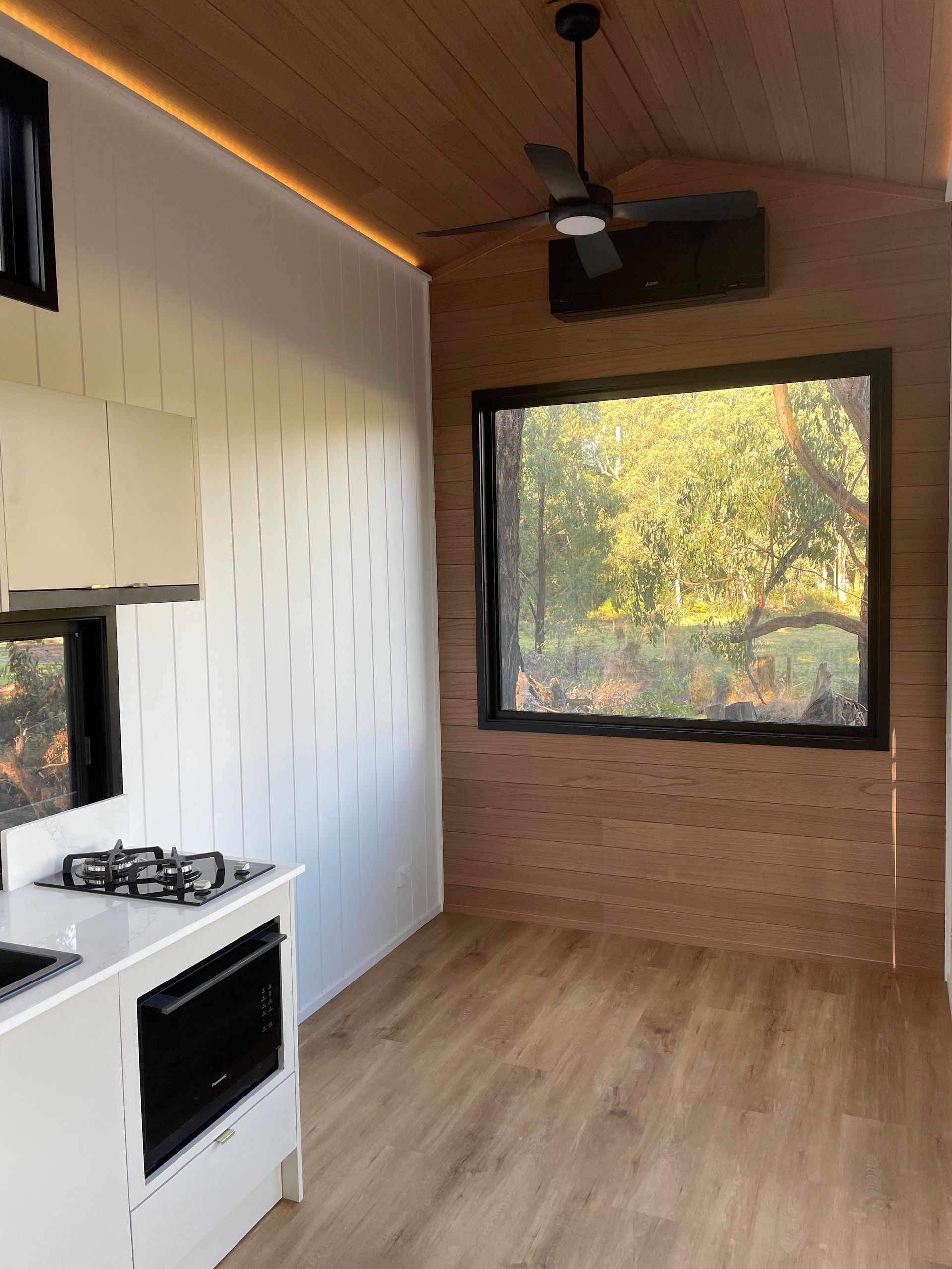 Interior of a tiny home with kitchen, window with a forest view, and ceiling fan. Wooden and white finishes.