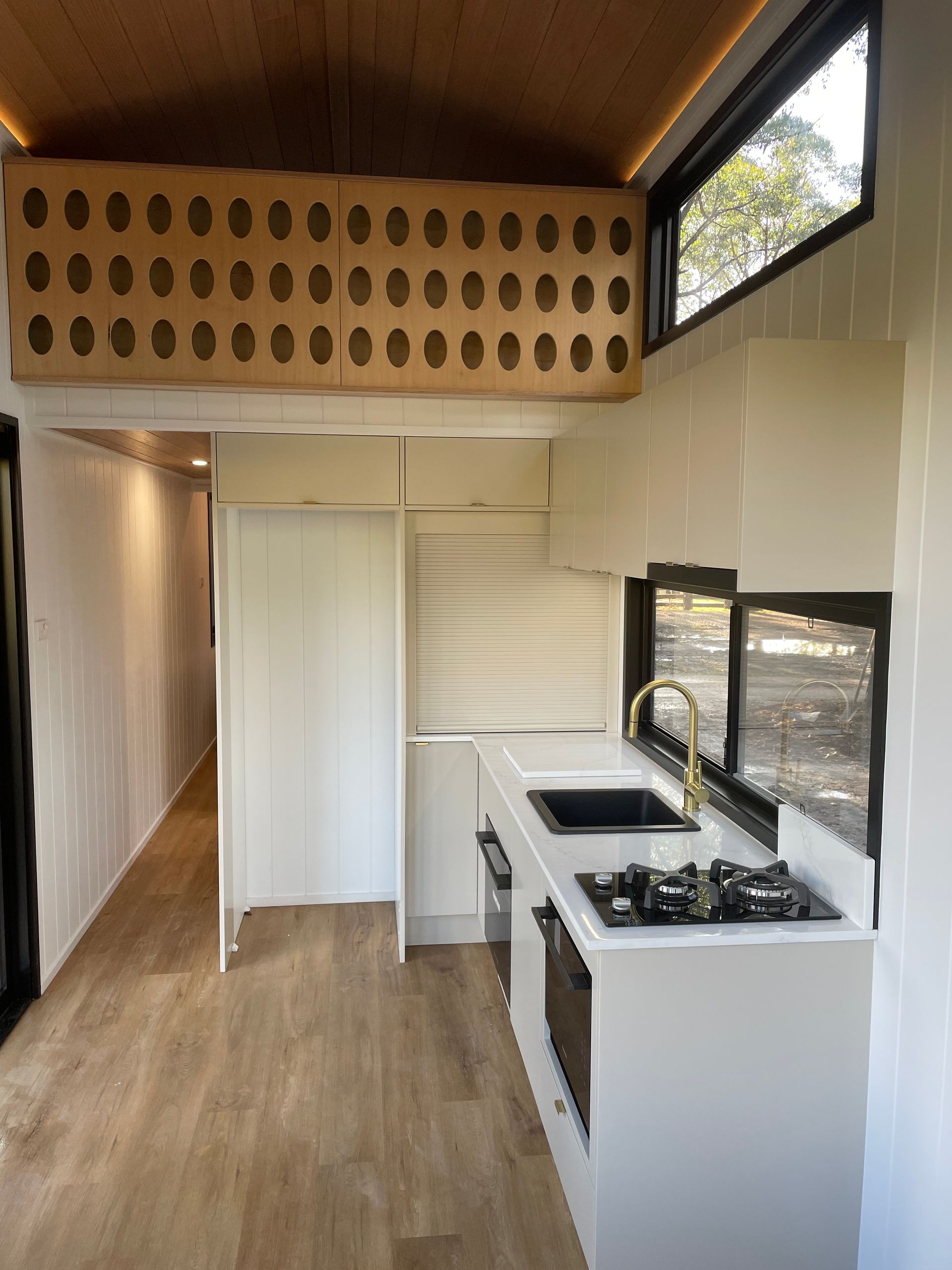 Interior of a tiny home with a white kitchen, black sink, and a wooden overhead storage unit.