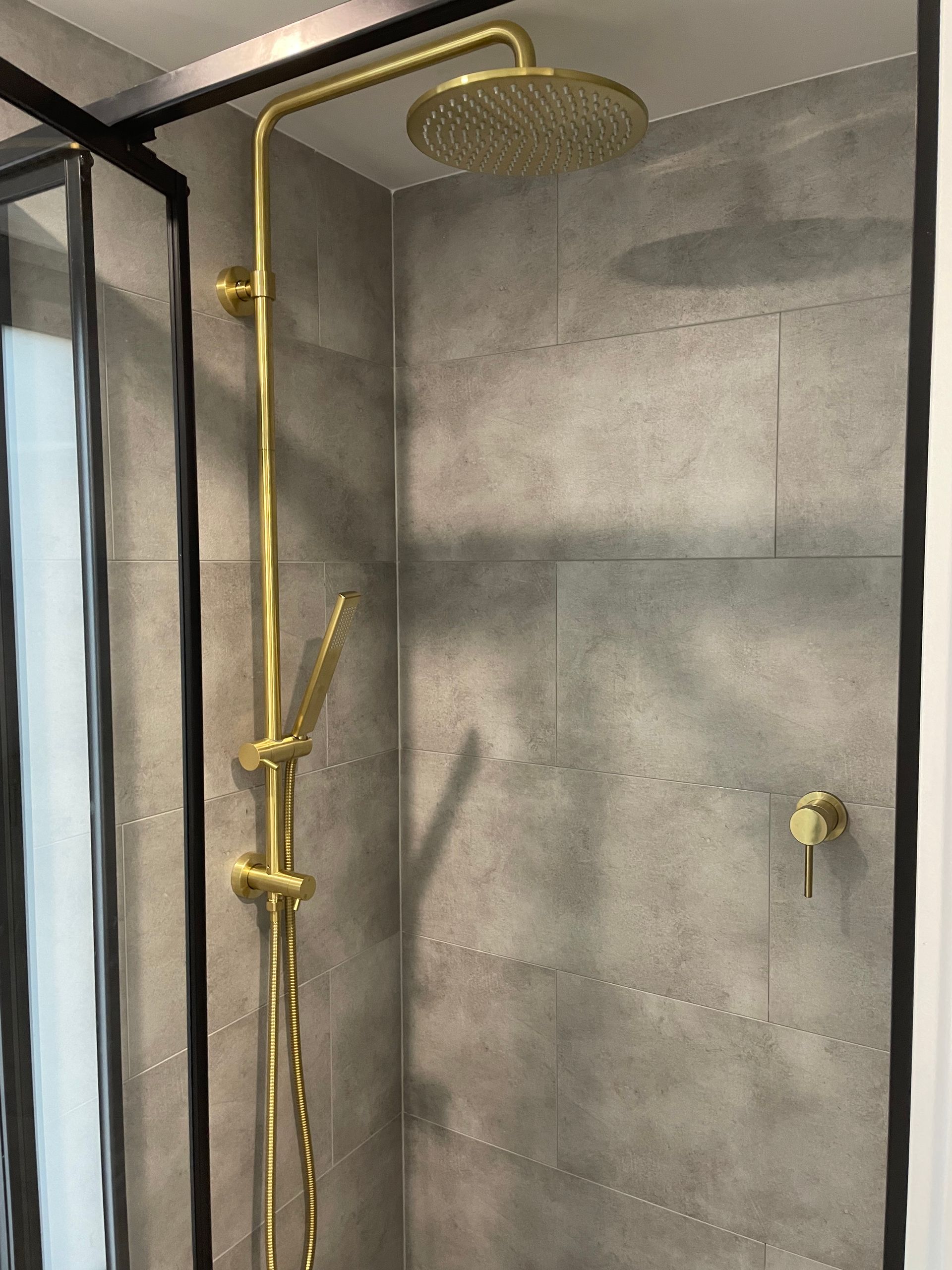 Gold showerhead and fixtures in a gray-tiled shower stall with a black-framed glass door.