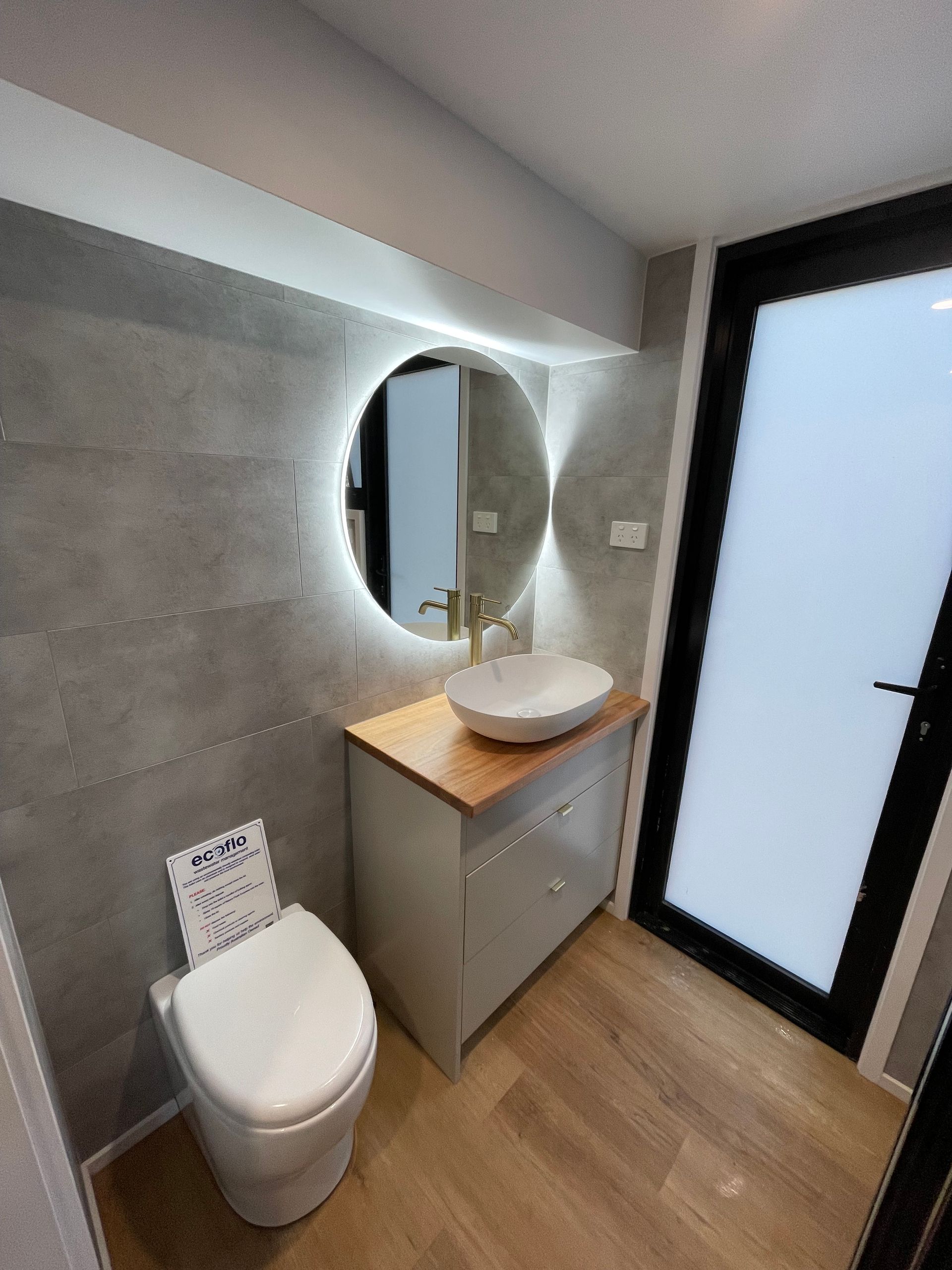 Bathroom with gray walls, wooden vanity, round mirror with lights, and frosted glass door.