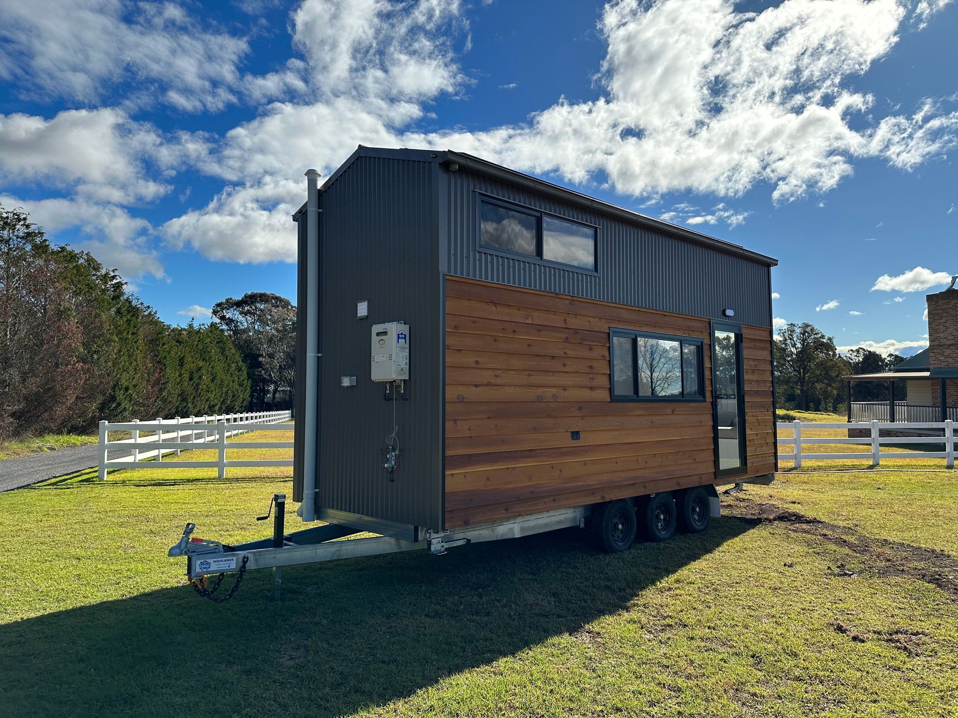 Tiny house on wheels, gray and brown exterior, on grass, blue sky background.