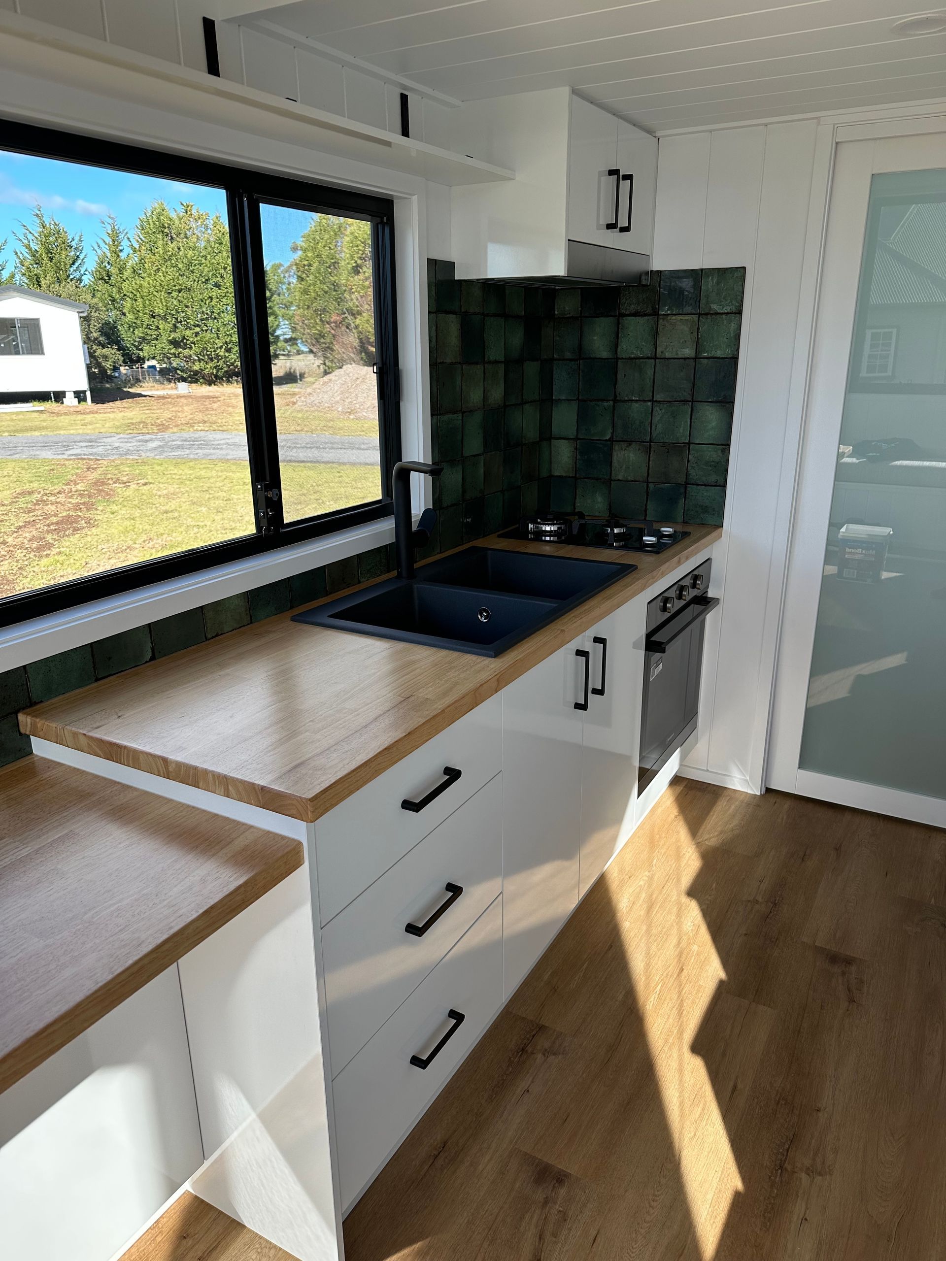 Modern kitchen with white cabinets, black countertop, and green tiled backsplash near a window.